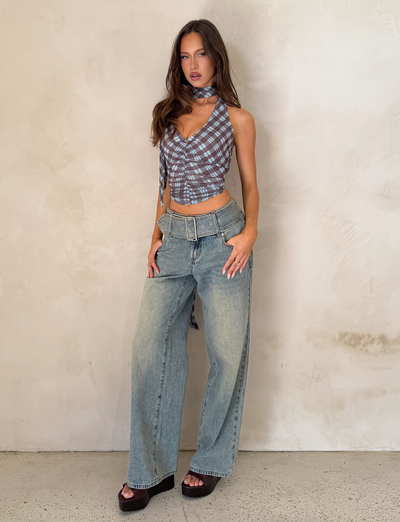 A woman stands against a light, textured wall wearing TIGERMIST’s REEF JEAN - LIGHTWASH with a detachable belt, a blue plaid crop top, black platform sandals, and a matching scarf around her neck.