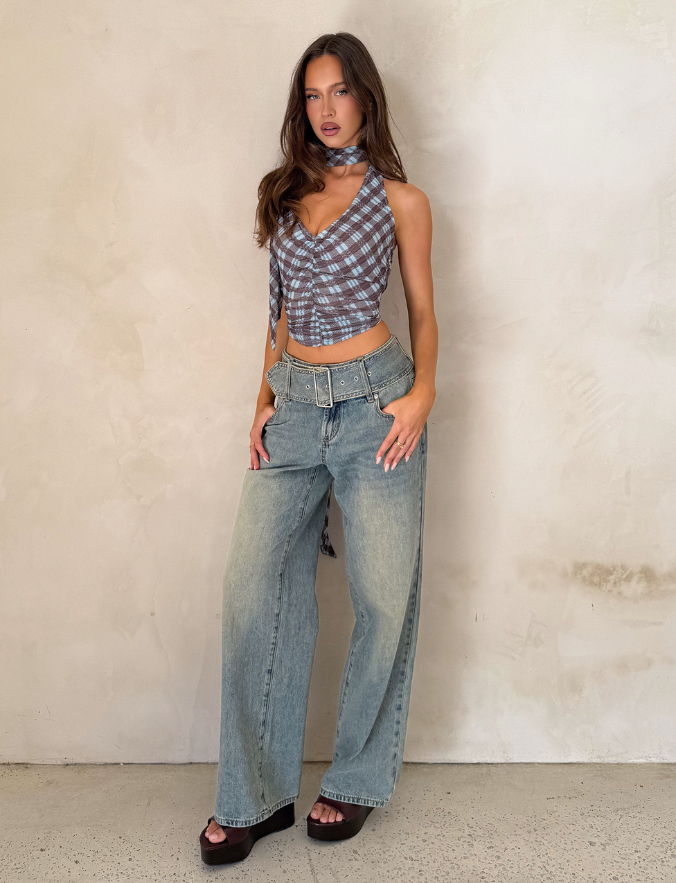 A woman stands against a light, textured wall wearing TIGERMIST’s REEF JEAN - LIGHTWASH with a detachable belt, a blue plaid crop top, black platform sandals, and a matching scarf around her neck.