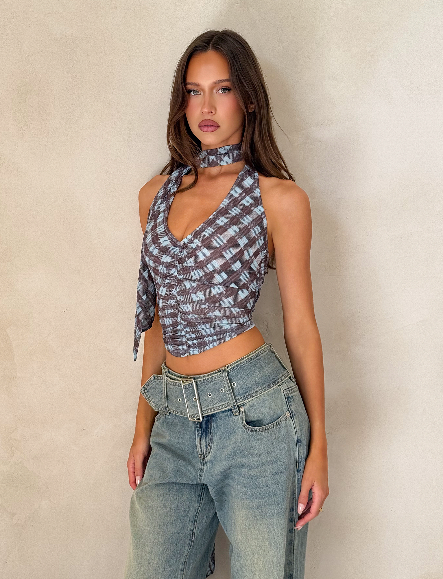A woman with long brown hair wears the TIGERMIST NOBLE TOP - BLUE CHECK, paired with a matching neck scarf and high-waisted, wide-leg jeans, standing against a light beige textured wall and gazing into the camera.