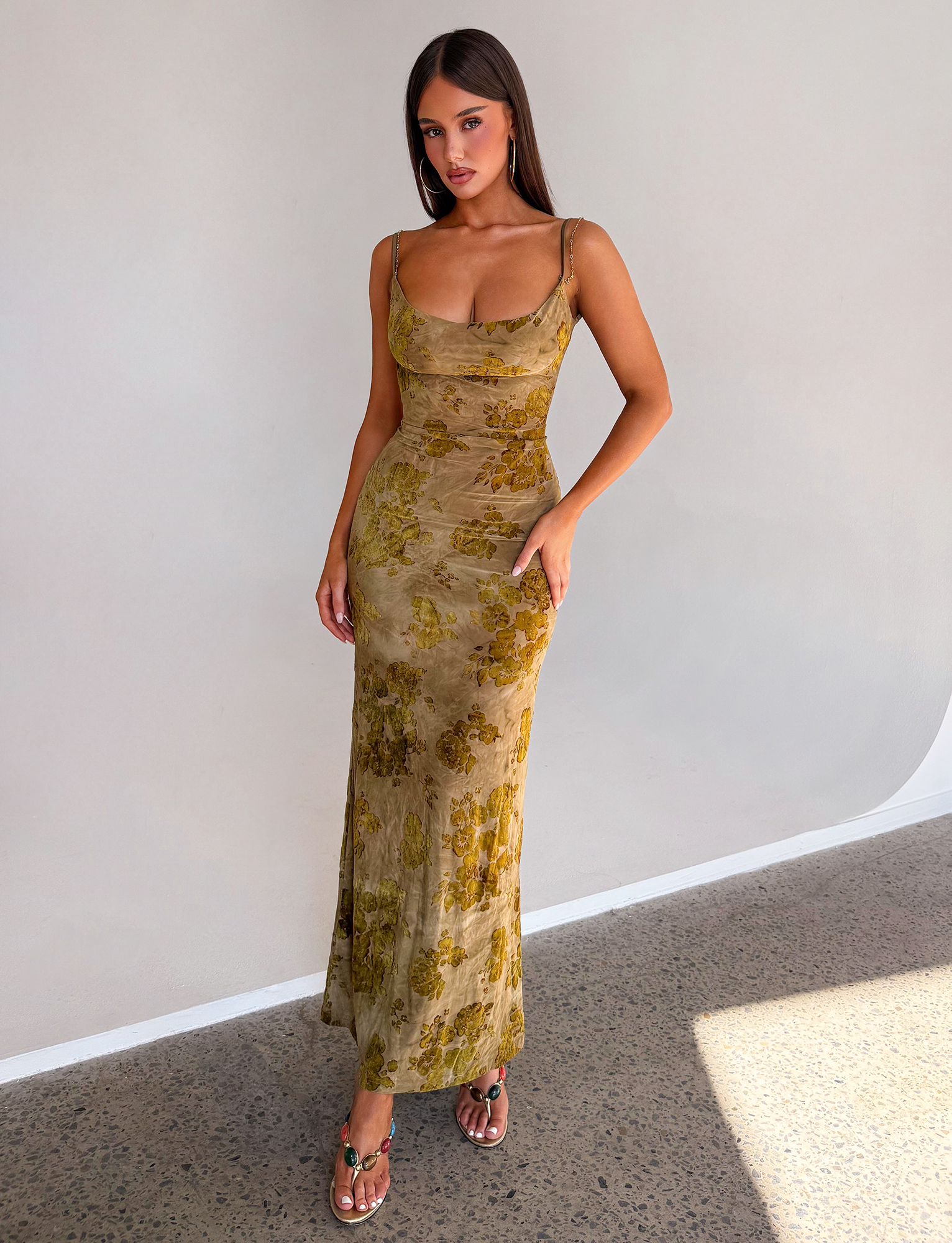 A woman with straight dark hair and natural makeup stands against a plain wall wearing the TIGERMIST SPARROW MAXI DRESS in green burnout—a fitted, sleeveless, floor-length floral dress—paired with sandals.