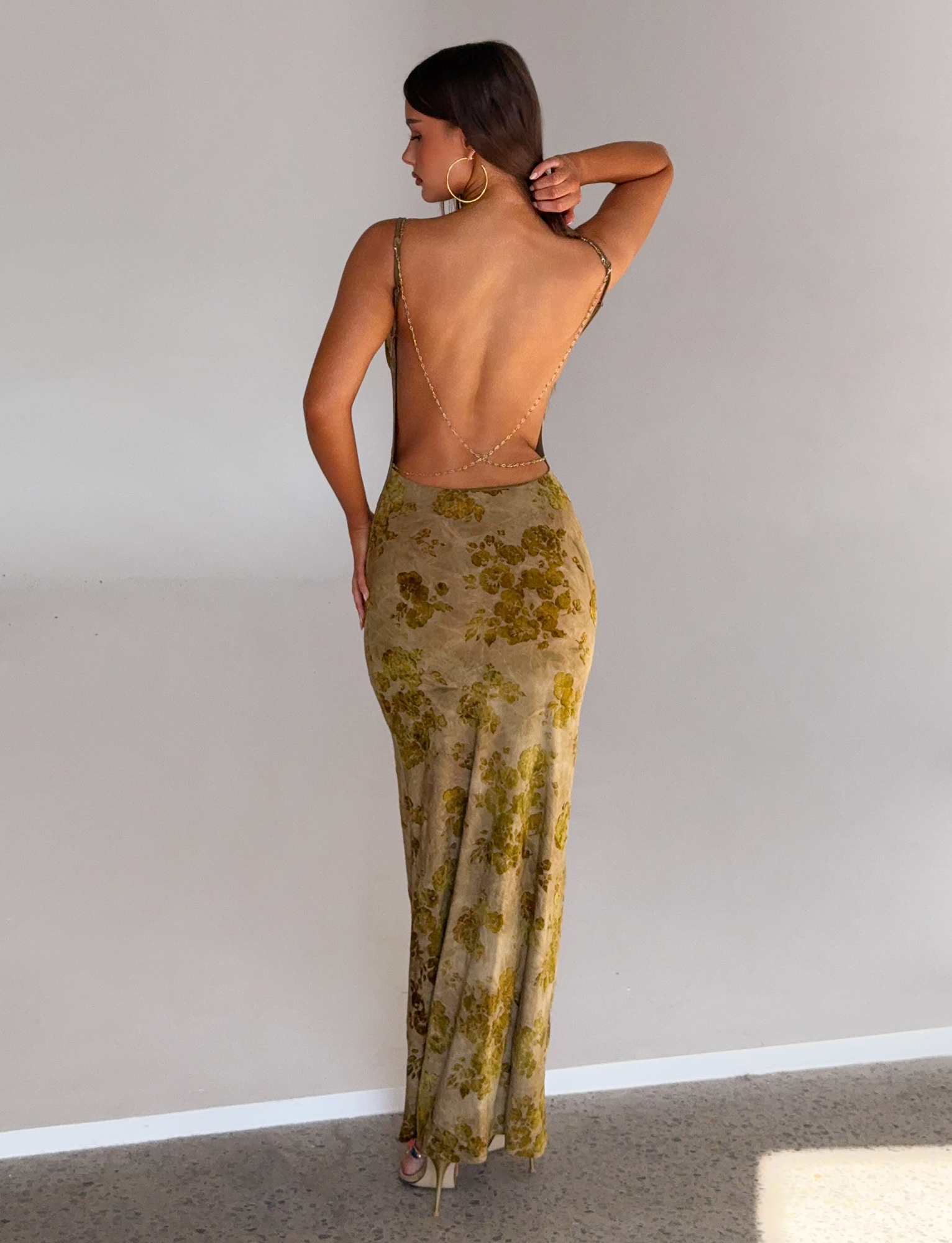 A woman faces away, wearing the TIGERMIST SPARROW MAXI DRESS in green burnout with gold chain straps and large hoop earrings, standing against a plain light-colored wall.