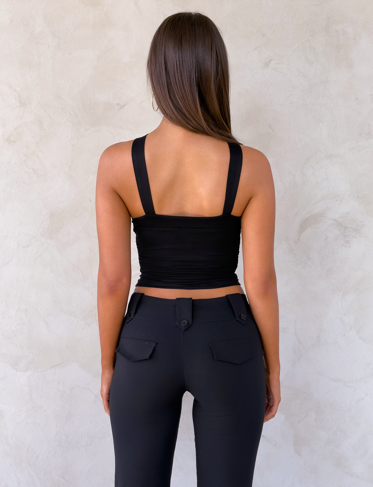 A woman with long brown hair faces a light-textured wall, wearing the TIGERMIST FINCH TOP in black and high-waisted black pants with pocket flaps and visible buttons.