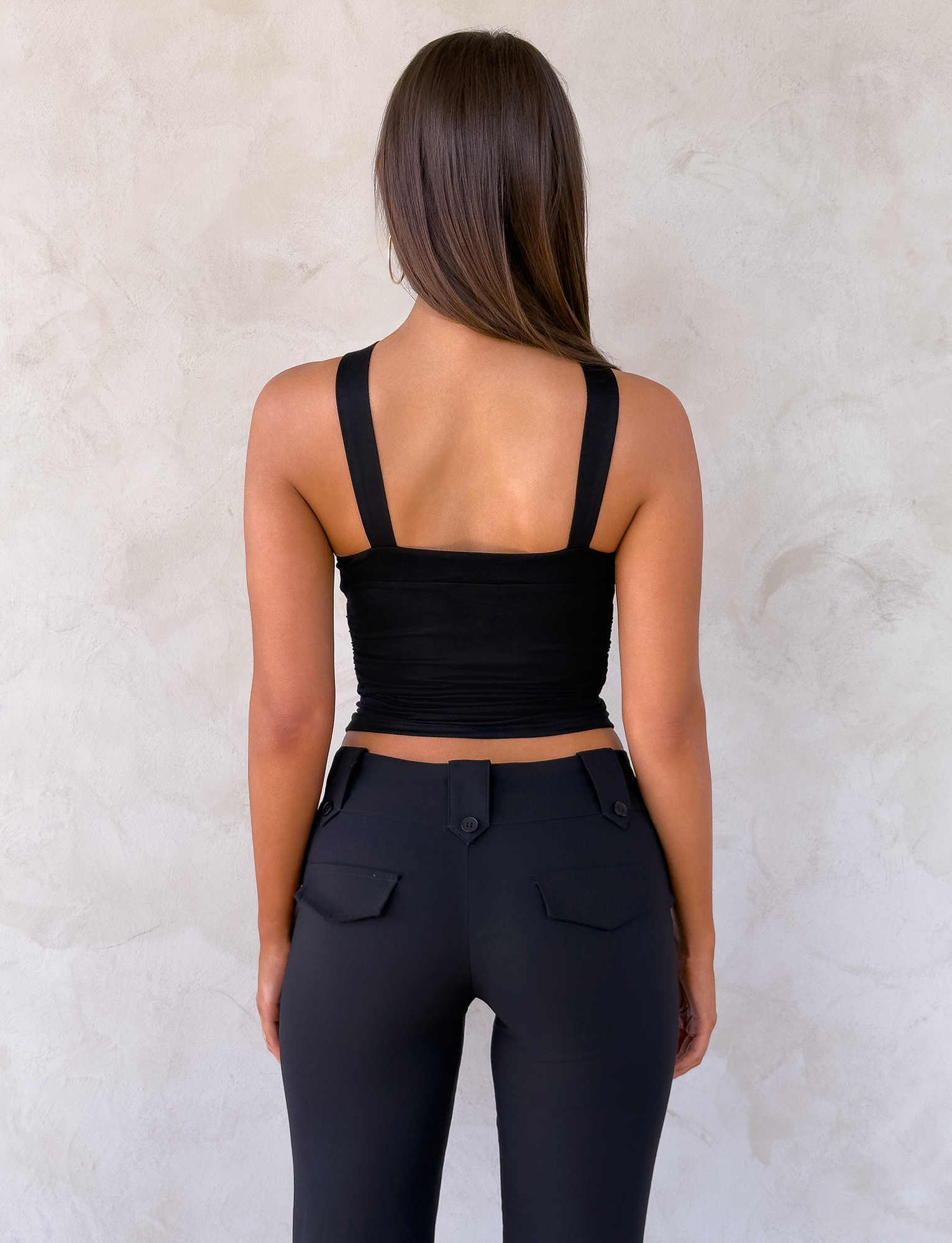 A woman with long brown hair faces a light-textured wall, wearing the TIGERMIST FINCH TOP in black and high-waisted black pants with pocket flaps and visible buttons.