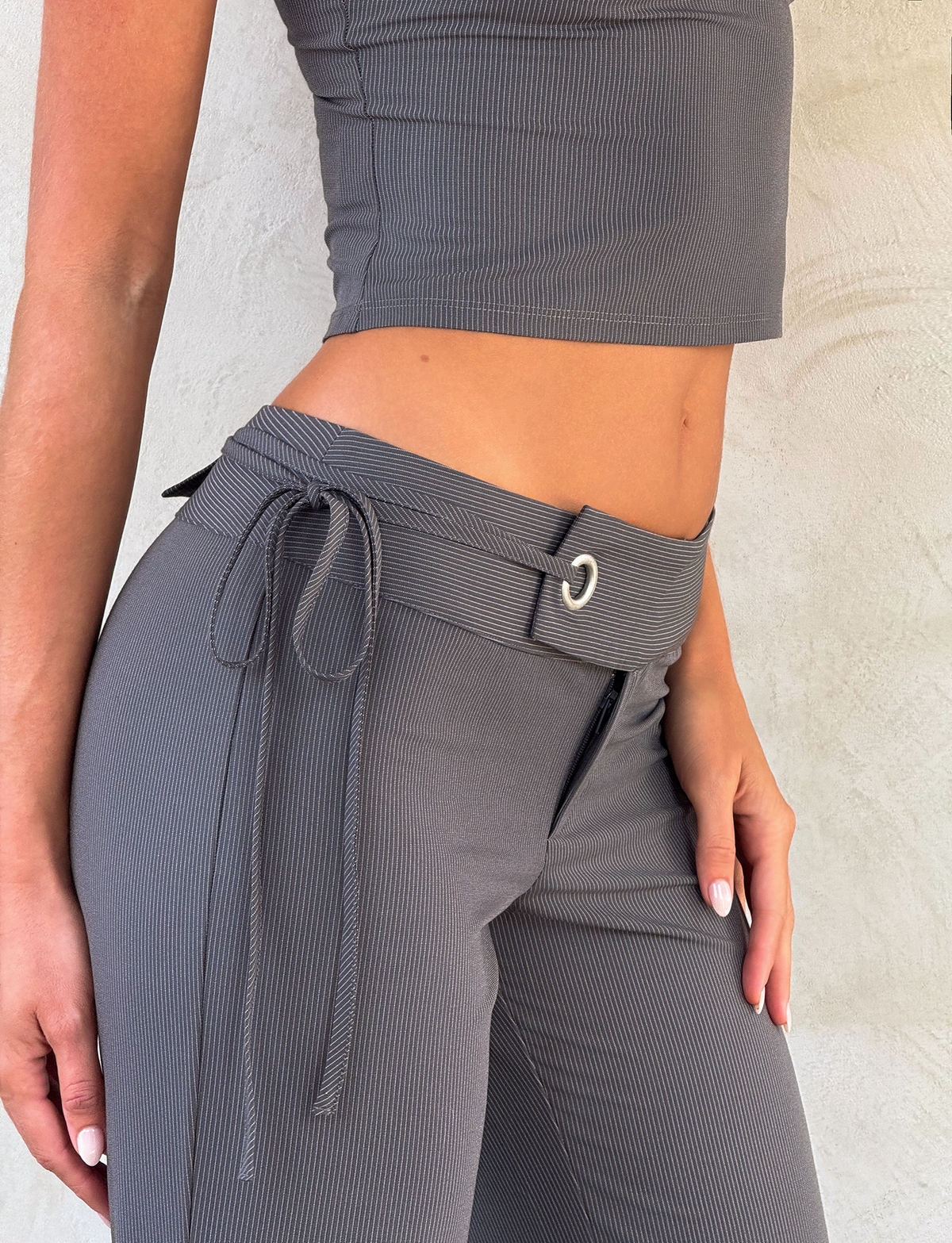 A person wearing the EMORY PANT - GREY PINSTRIPE by TIGERMIST, featuring a wide belt and tie detail, paired with a ribbed grey crop top and standing against a light textured wall.