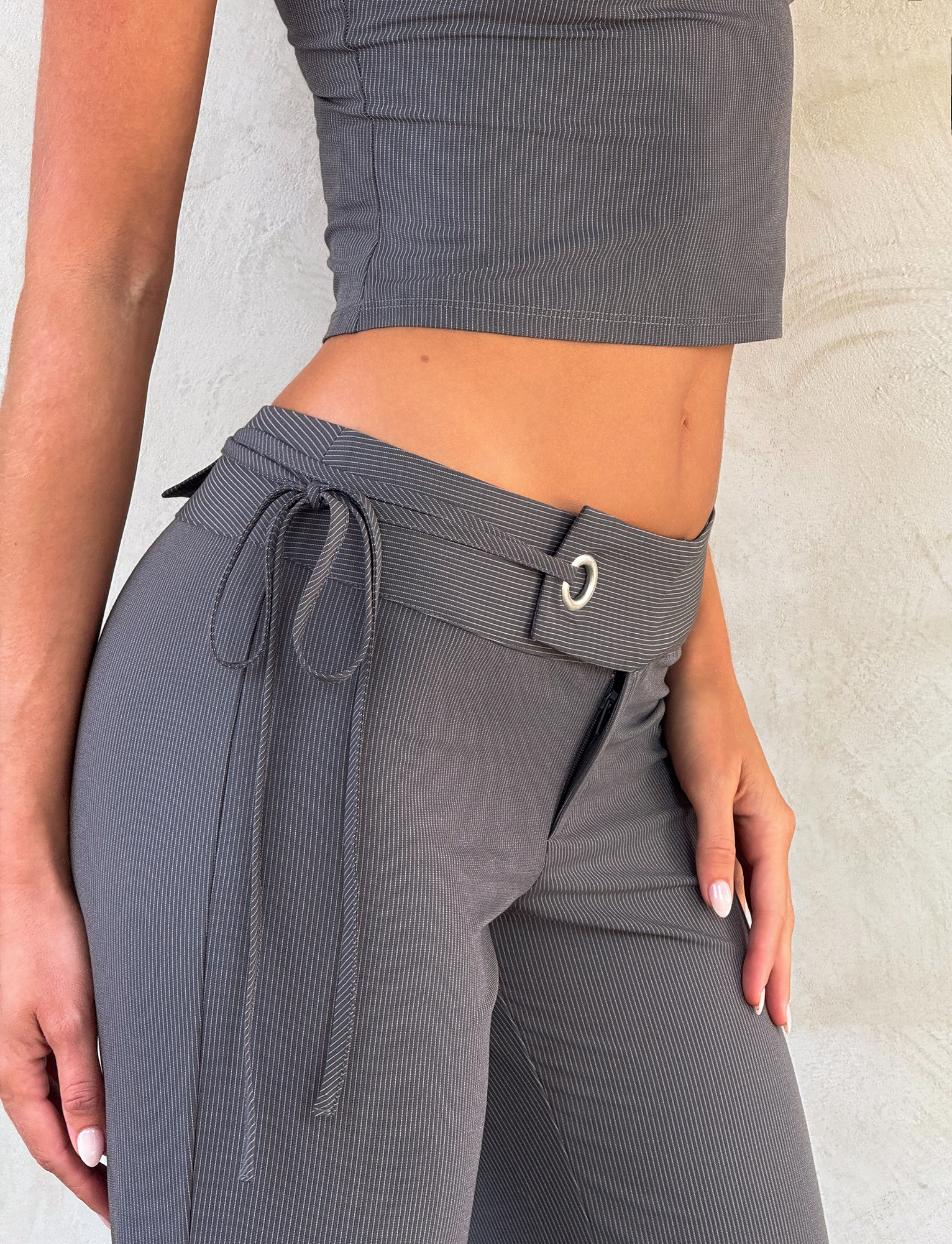 A person wearing the EMORY PANT - GREY PINSTRIPE by TIGERMIST, featuring a wide belt and tie detail, paired with a ribbed grey crop top and standing against a light textured wall.