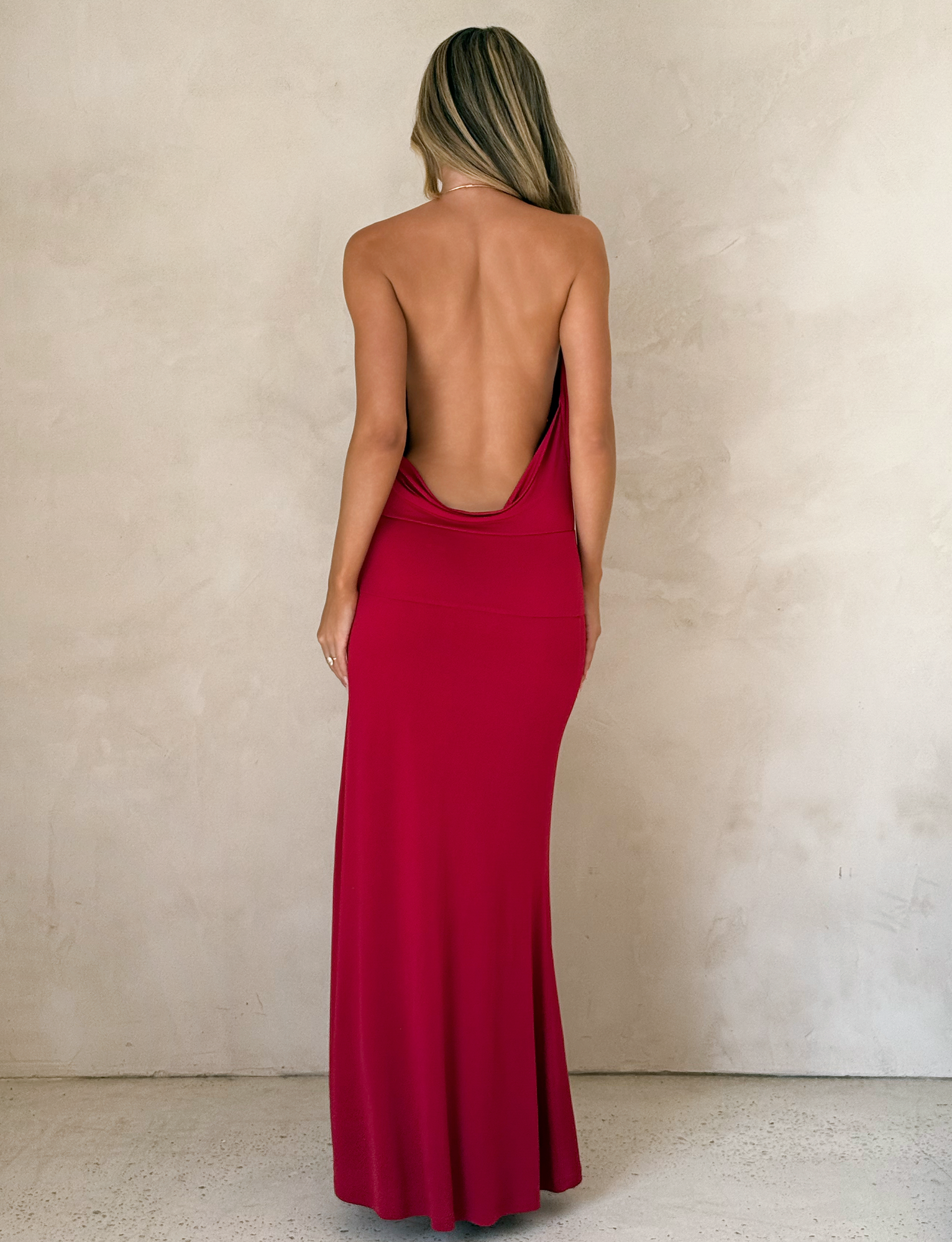 A woman with long blonde hair, facing away from the camera, wears the TIGERMIST TARRIN MAXI DRESS in red. The dress features a cowl neckline and thigh-high slit. She stands indoors against a light-textured wall.