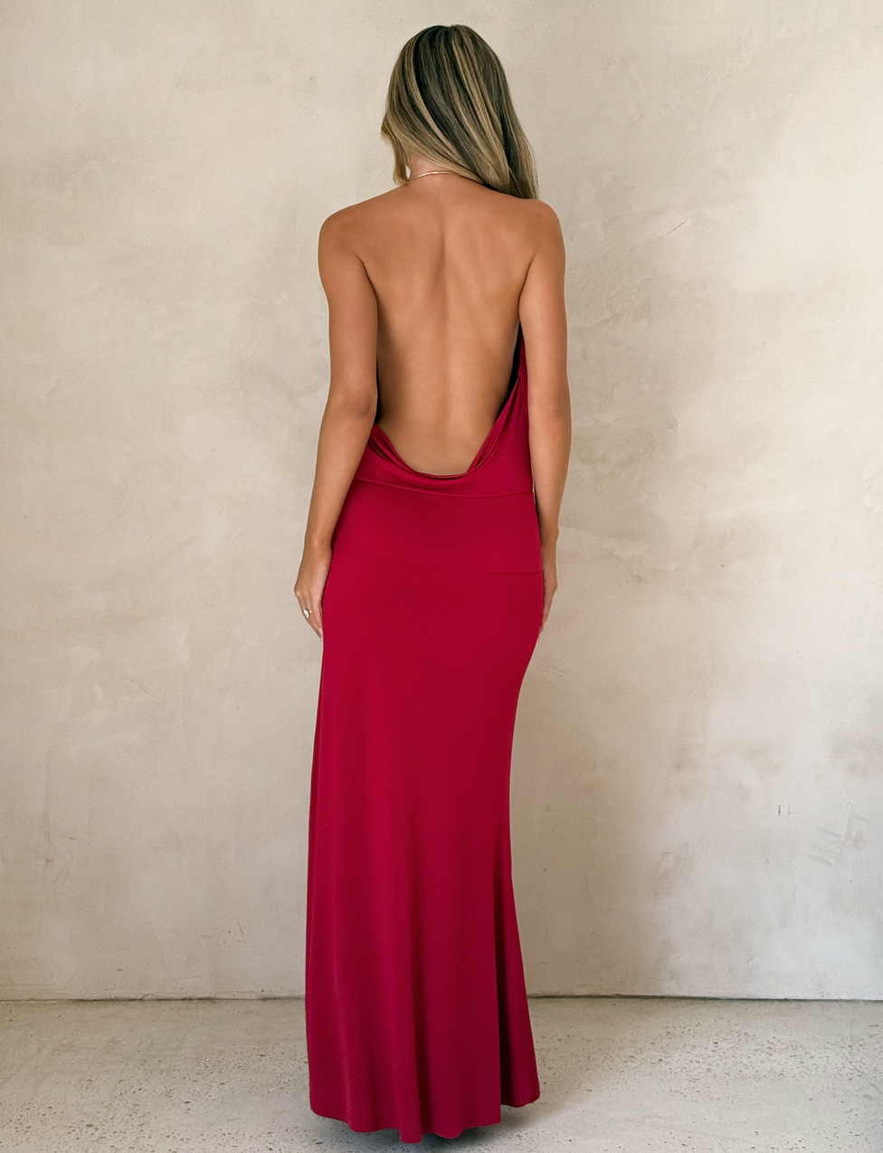 A woman with long blonde hair, facing away from the camera, wears the TIGERMIST TARRIN MAXI DRESS in red. The dress features a cowl neckline and thigh-high slit. She stands indoors against a light-textured wall.