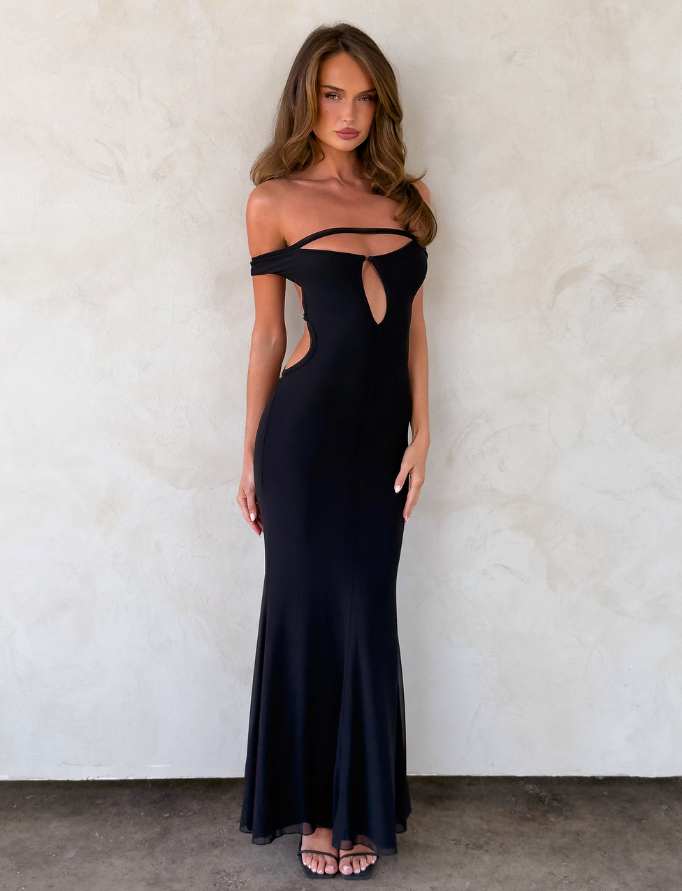 A woman stands against a light textured wall wearing the TIGERMIST KERRIGAN MAXI DRESS in black, featuring off-shoulder sleeves and chest and waist cutouts. Her fitted silhouette is complemented by long, wavy brown hair and open-toed heels.