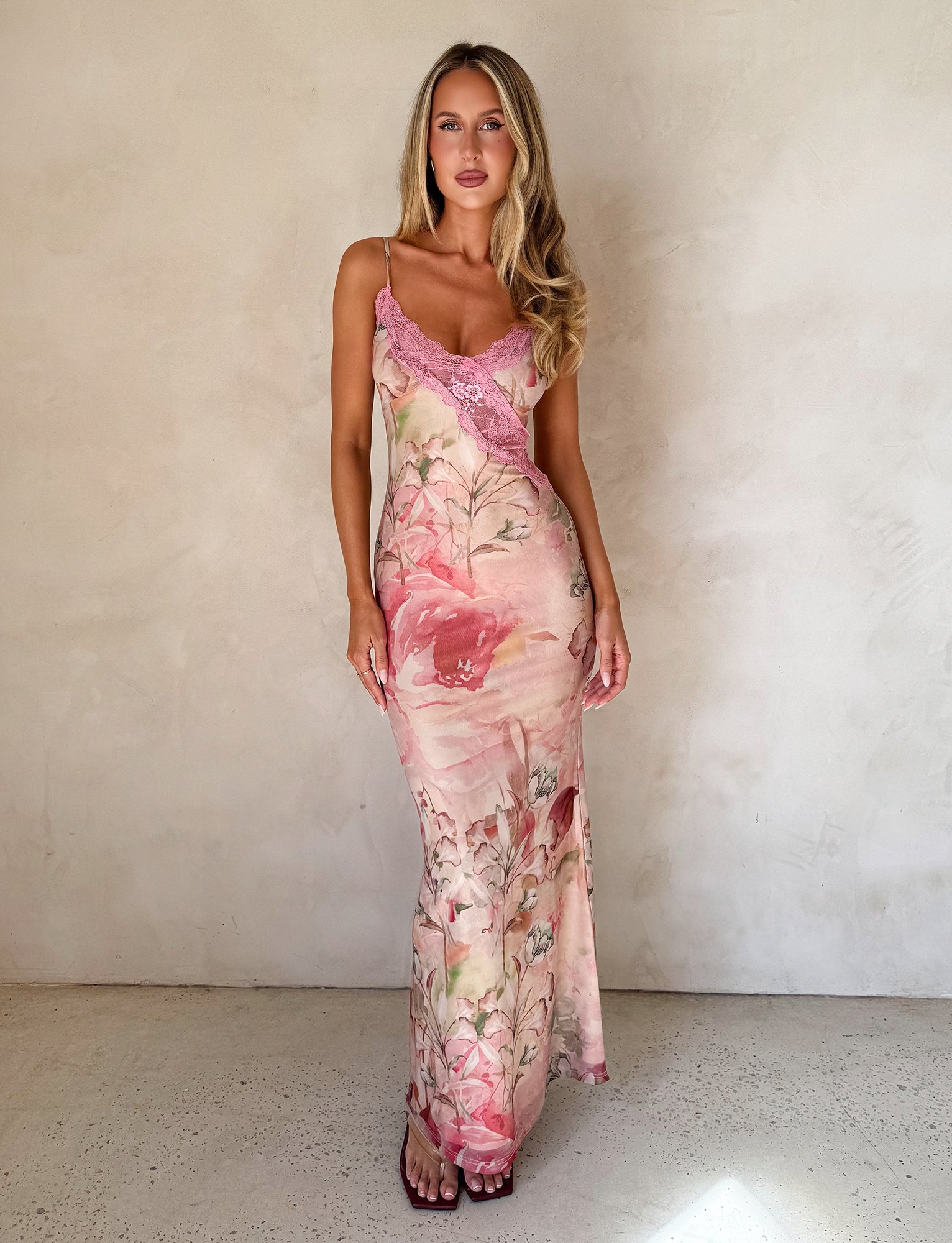 A woman with long, wavy blonde hair stands against a light textured wall wearing the TIGERMIST LYRE MAXI DRESS in Watercolour Floral, featuring sheer pink lace details and dark sandals, exuding relaxed confidence.