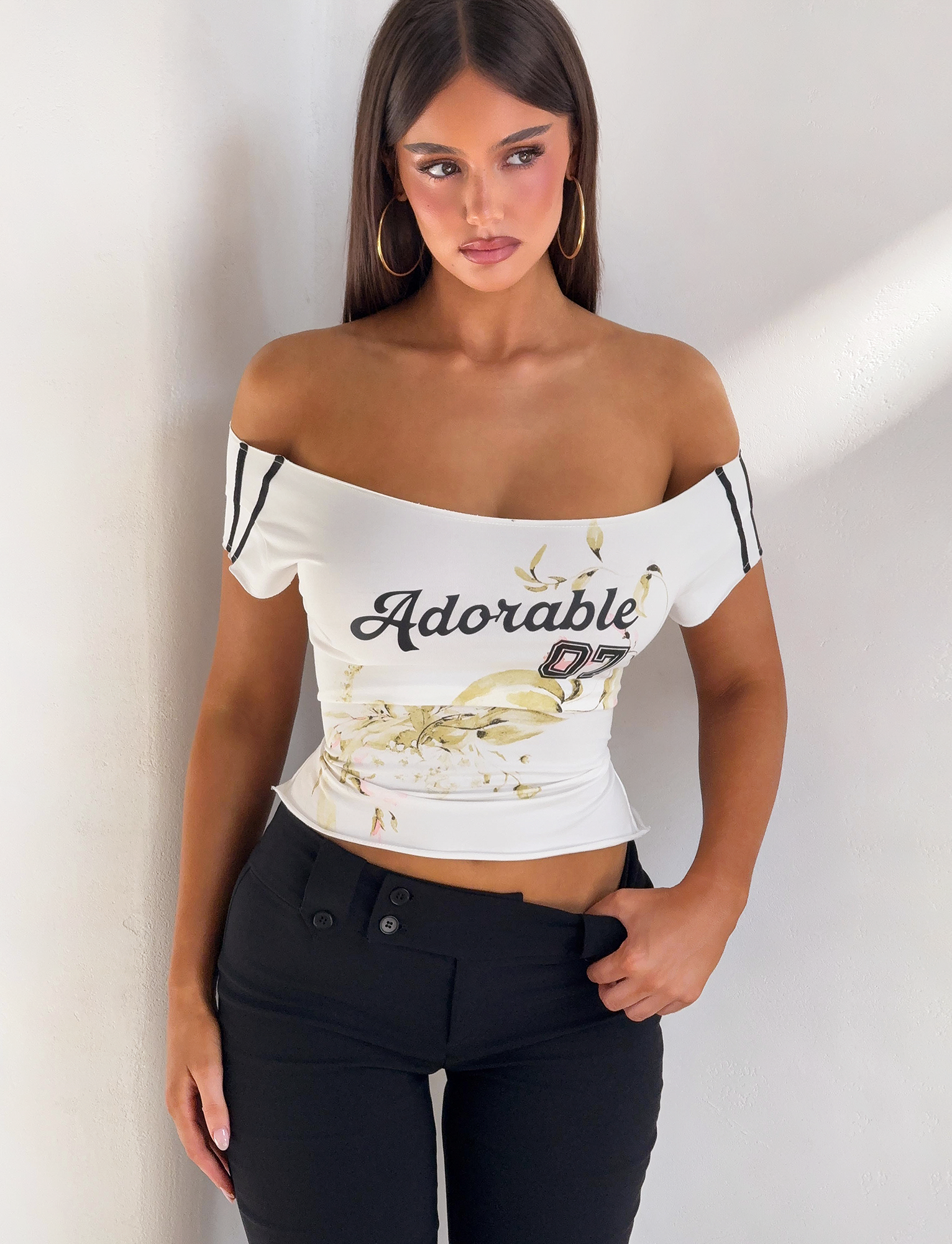 A woman with long brown hair stands against a white wall wearing the TIGERMIST FYNN TEE - WHITE, an off-shoulder top with “Adorable 07” and floral graphic, paired with black high-waisted pants. She gazes to the side, expression neutral.
