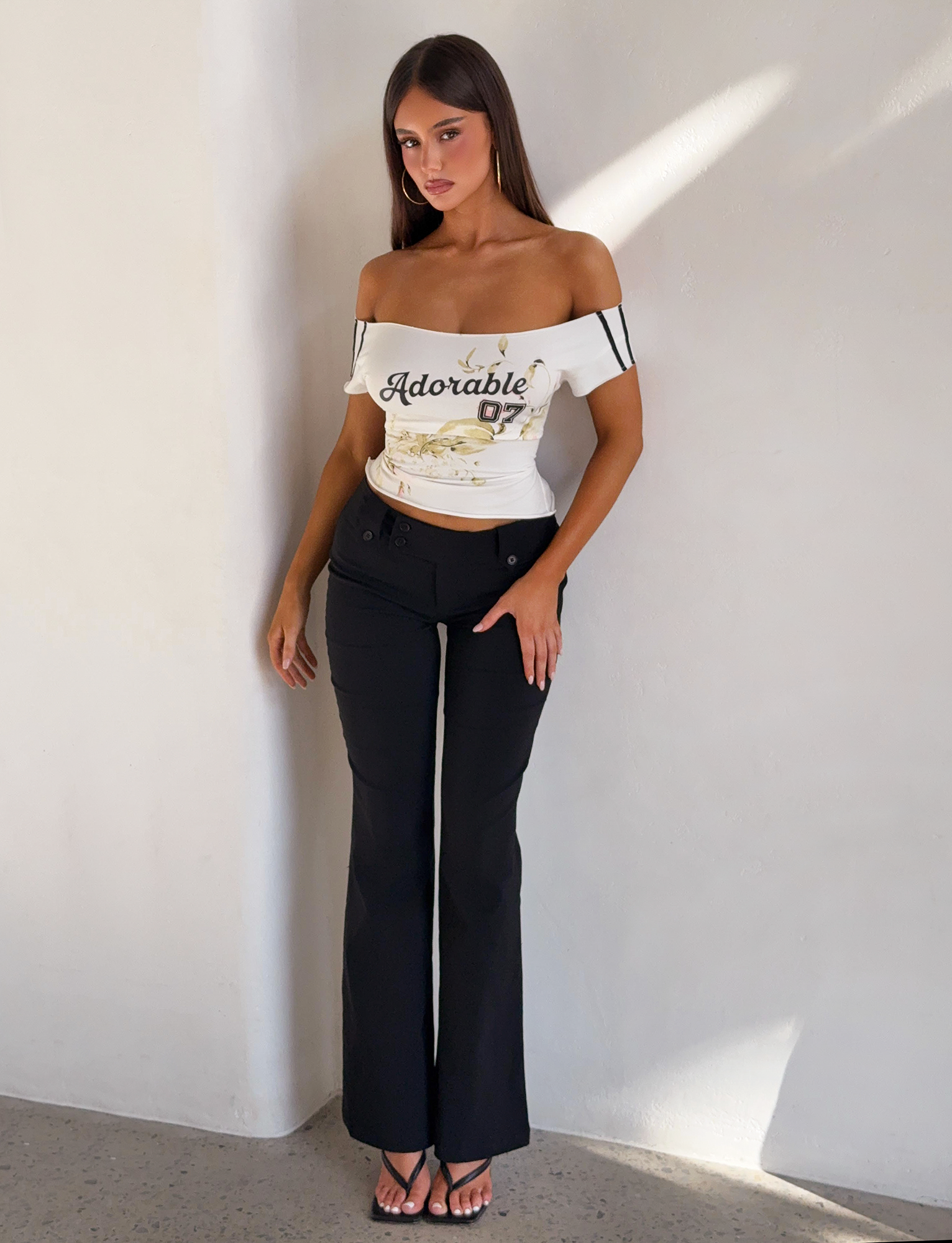 A woman stands indoors against a white wall, wearing the TIGERMIST FYNN TEE - WHITE with "Adorable 07" print, paired with black pants and sandals. She has long straight hair and a neutral expression.