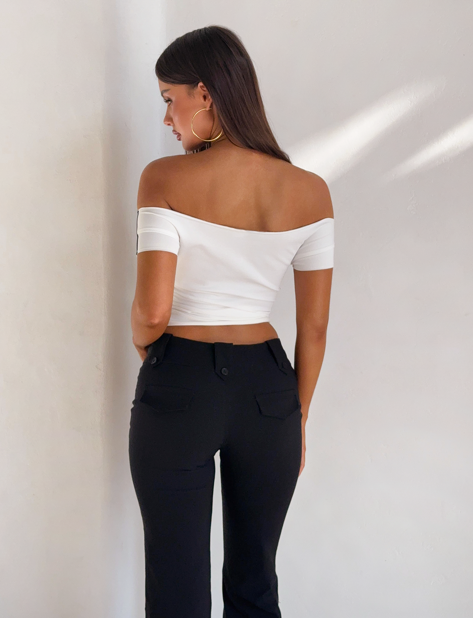 A woman with long brown hair stands facing a white wall, wearing the TIGERMIST FYNN TEE in white off-shoulder style, paired with black pants and large hoop earrings. She is turned slightly to the side, showing her back and shoulders.