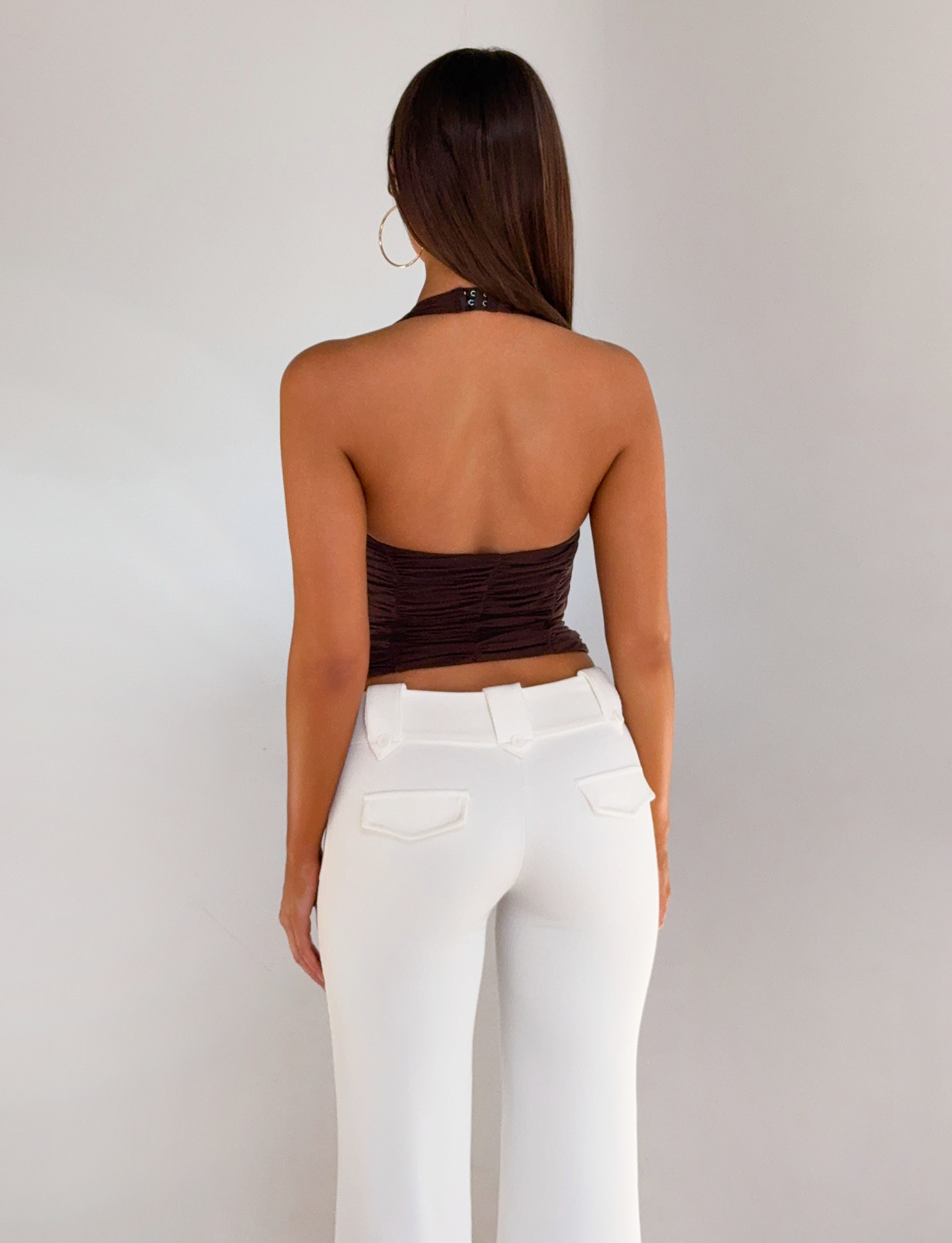 A woman with long brown hair faces away, wearing the TIGERMIST BRIALLEN TOP in chocolate and high-waisted white pants with back pockets, set against a plain light background.