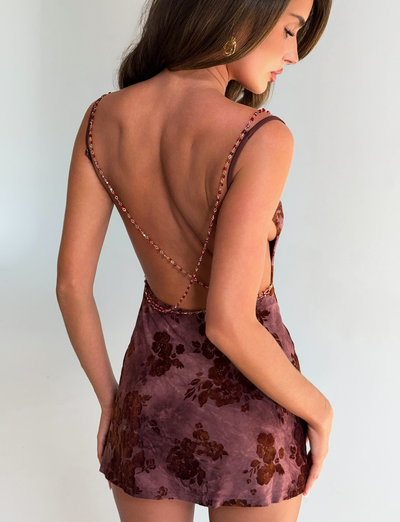 A woman with long brown hair wears the TIGERMIST SPARROW MINI DRESS in Chocolate Burnout, standing with her back to the camera and glancing over her shoulder.