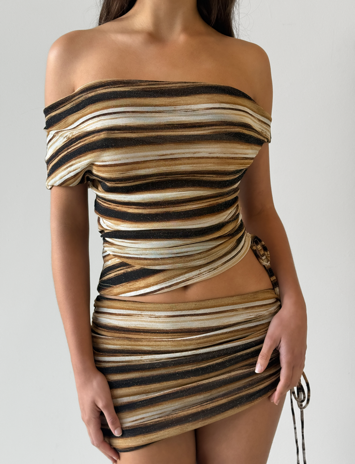 A woman wears the TIGERMIST BRAELYN TOP in brown stripe—an off-the-shoulder, fitted, lurex two-piece with horizontal brown, gold, black, and white stripes. The wrapped short top and matching high-waisted mini skirt create a chic look.
