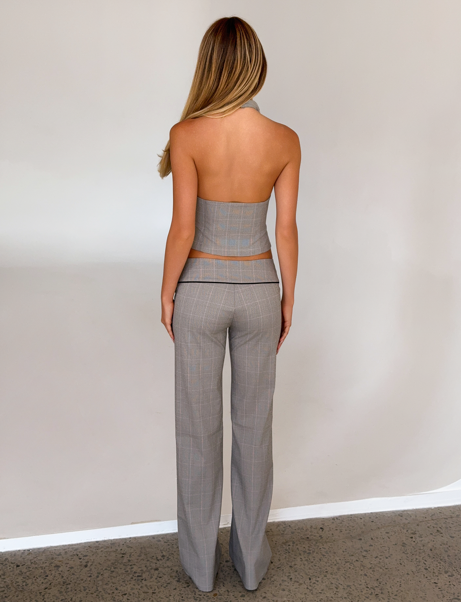 A woman with long blonde hair stands facing a light wall, wearing TIGERMIST’s TERESIA PANT in grey check with a matching strapless top. Her arms rest at her sides and her back is to the camera.