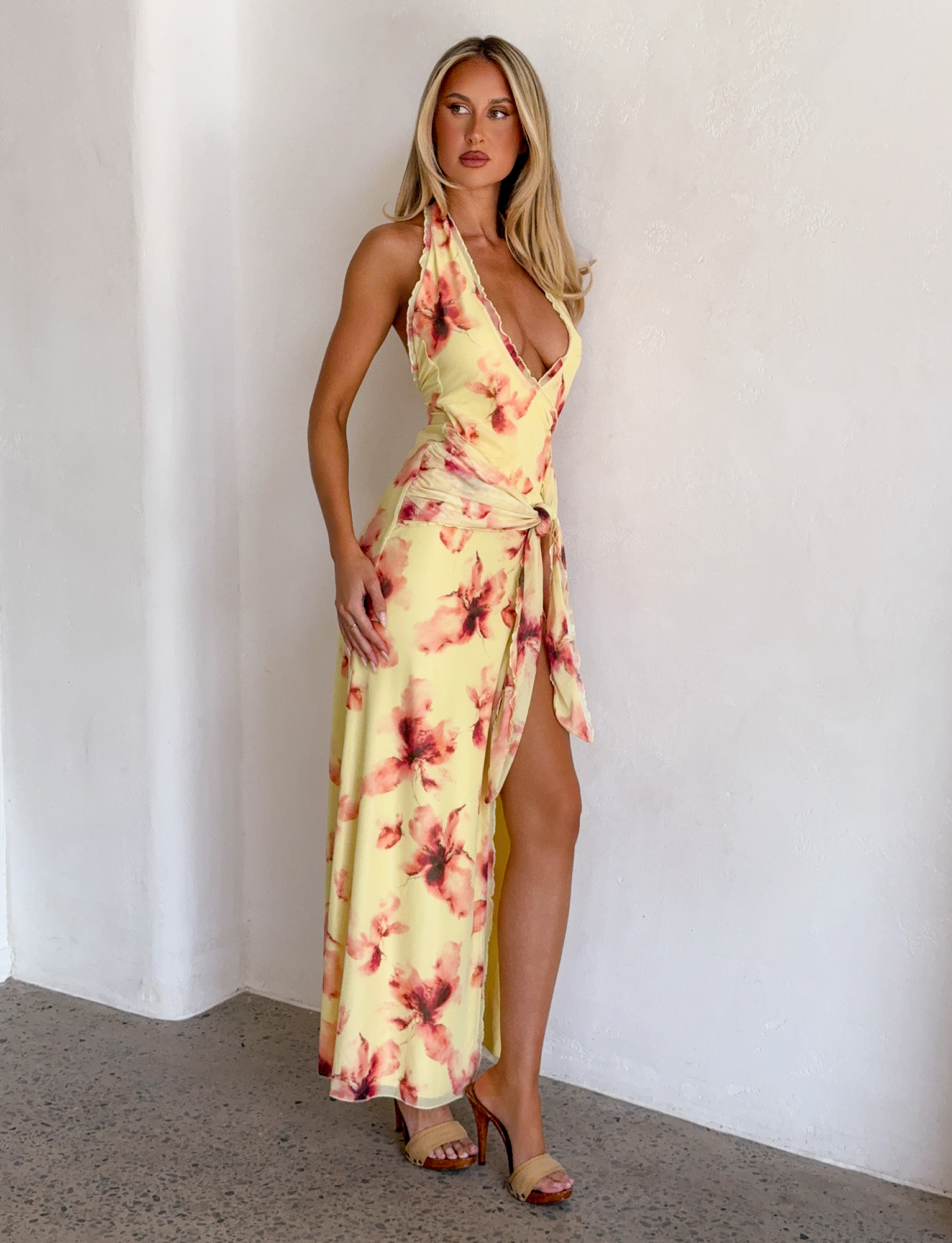 A woman poses indoors in the TIGERMIST ALLEGRA MAXI DRESS in yellow watercolour floral, featuring a sleeveless design, deep V-neckline and high leg split. She has long blonde hair and pairs the look with chic heeled sandals.