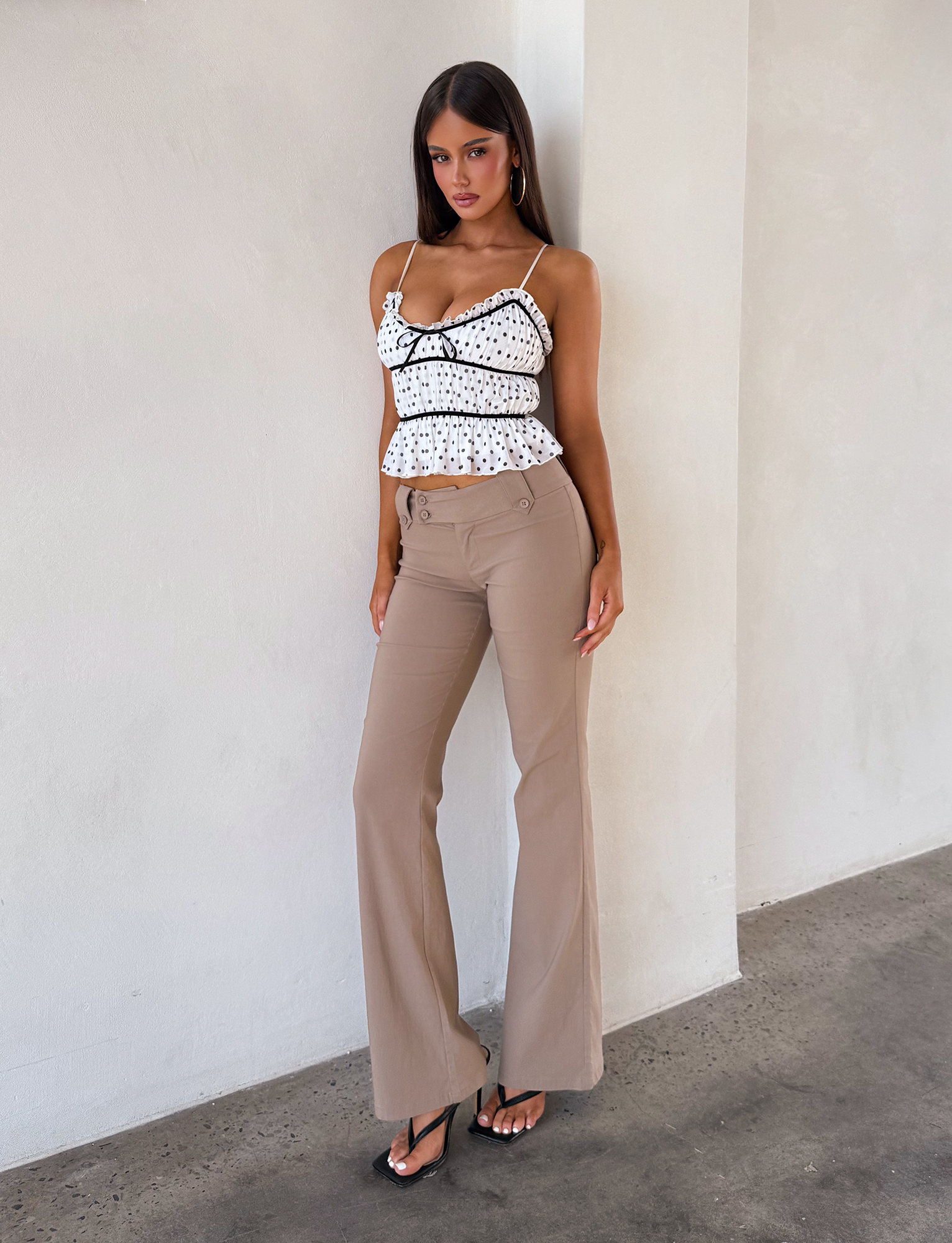 A woman stands against a light-colored wall, wearing the TIGERMIST LIORA TOP - POLKA DOT with adjustable straps and black trim, paired with beige high-waisted flared pants and black heeled sandals.
