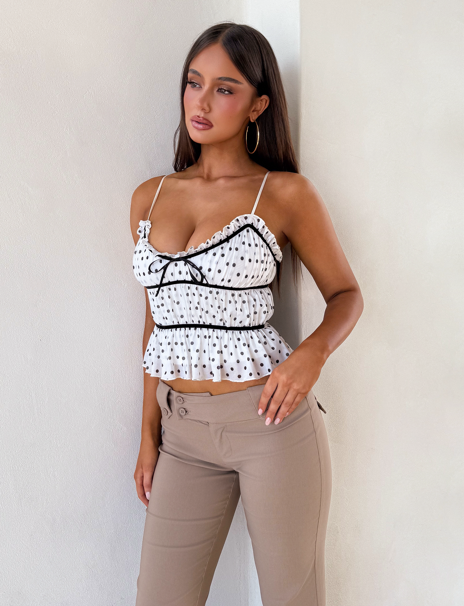 A woman with long dark hair and hoop earrings wears the TIGERMIST LIORA TOP - POLKA DOT cami and beige high-waisted pants, standing against a light wall and looking to the side.