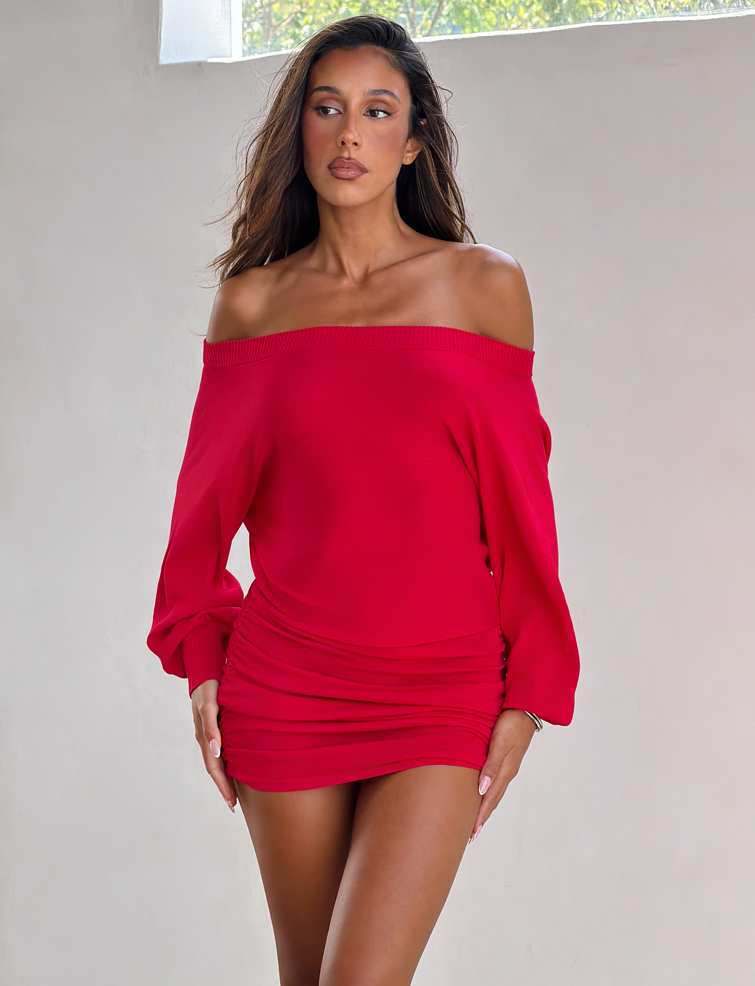 A woman with long brown hair wears the TIGERMIST JANINE MINI DRESS in red, featuring an off-shoulder design, long sleeves, and a ruched waist, standing indoors by a light-colored wall as natural light shines in.