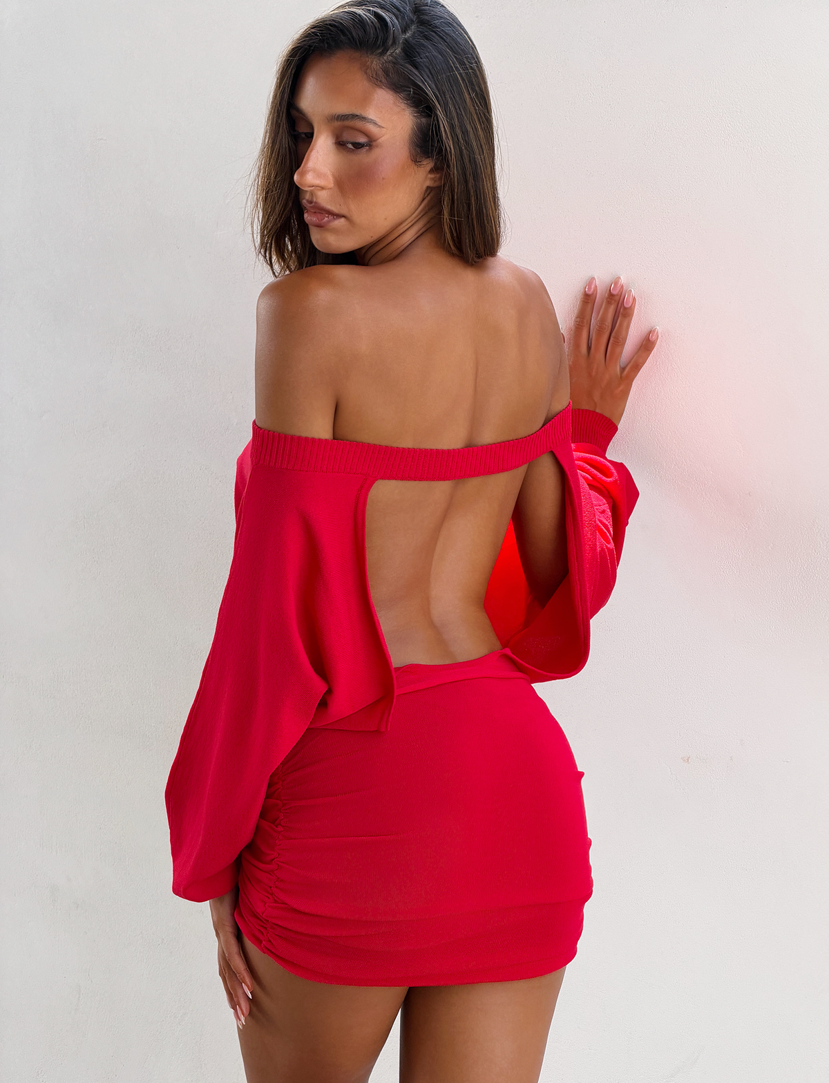 A woman stands against a white wall wearing the TIGERMIST JANINE MINI DRESS in red, featuring an off-shoulder neckline, open back, and ruched skirt, as she looks down over her shoulder.
