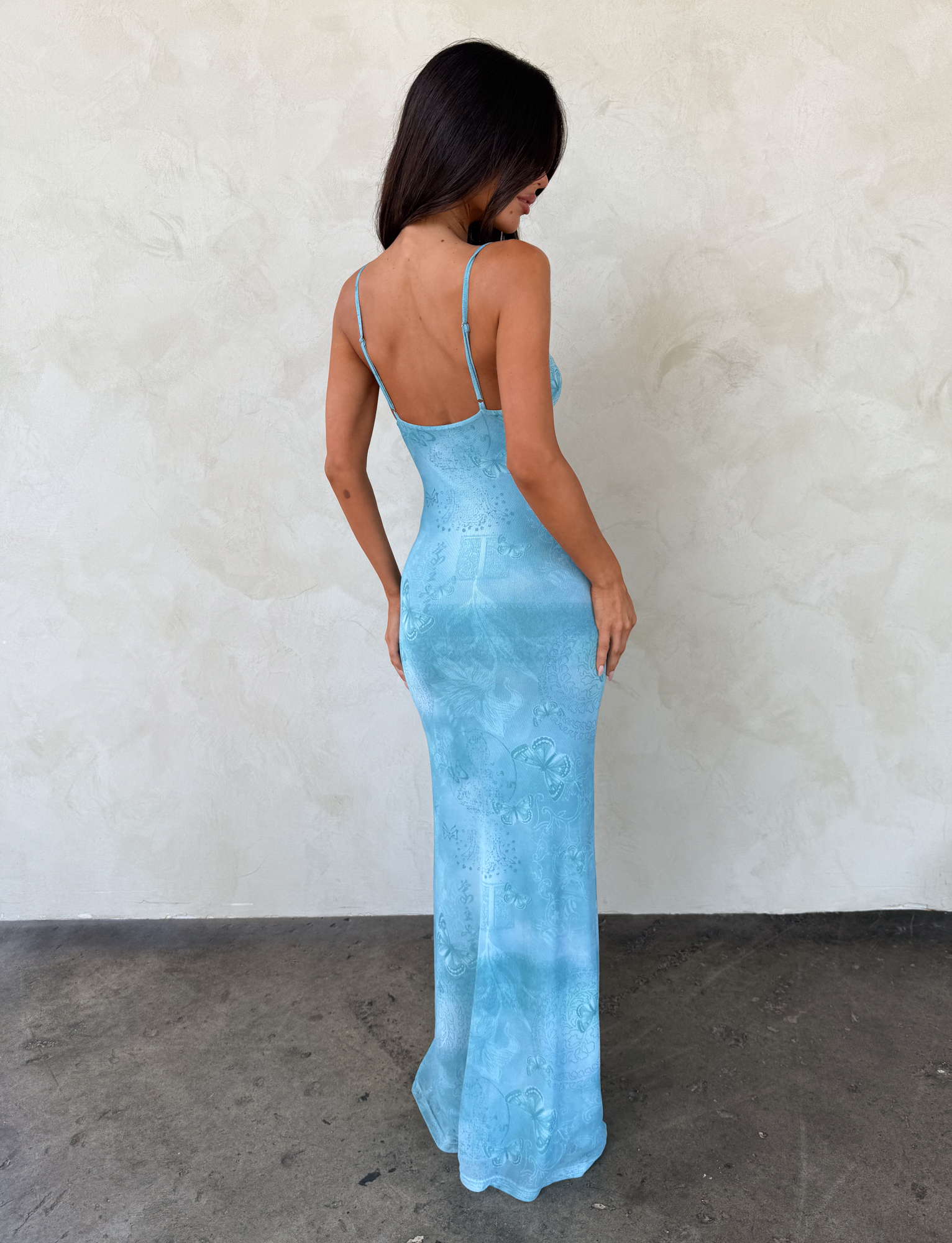 A woman with long dark hair wears the TIGERMIST GARNET MAXI DRESS in Teal Butterfly Paisley. She stands on a concrete floor, facing away from the camera, against a textured pale wall.