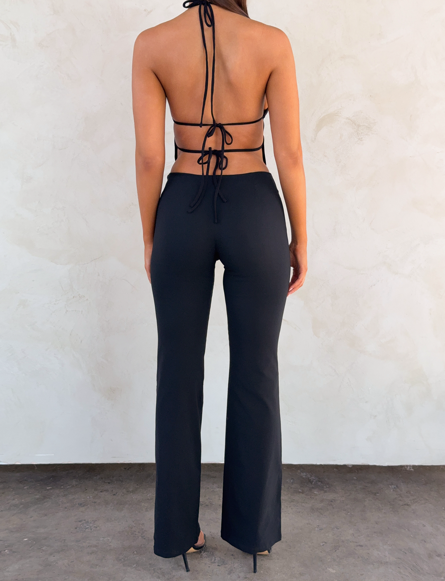 A woman faces a light wall, wearing TIGERMIST’s MARREN PANT in black, styled with a black halter top featuring an open back and tie details, plus black heels. Her arms hang relaxed at her sides.