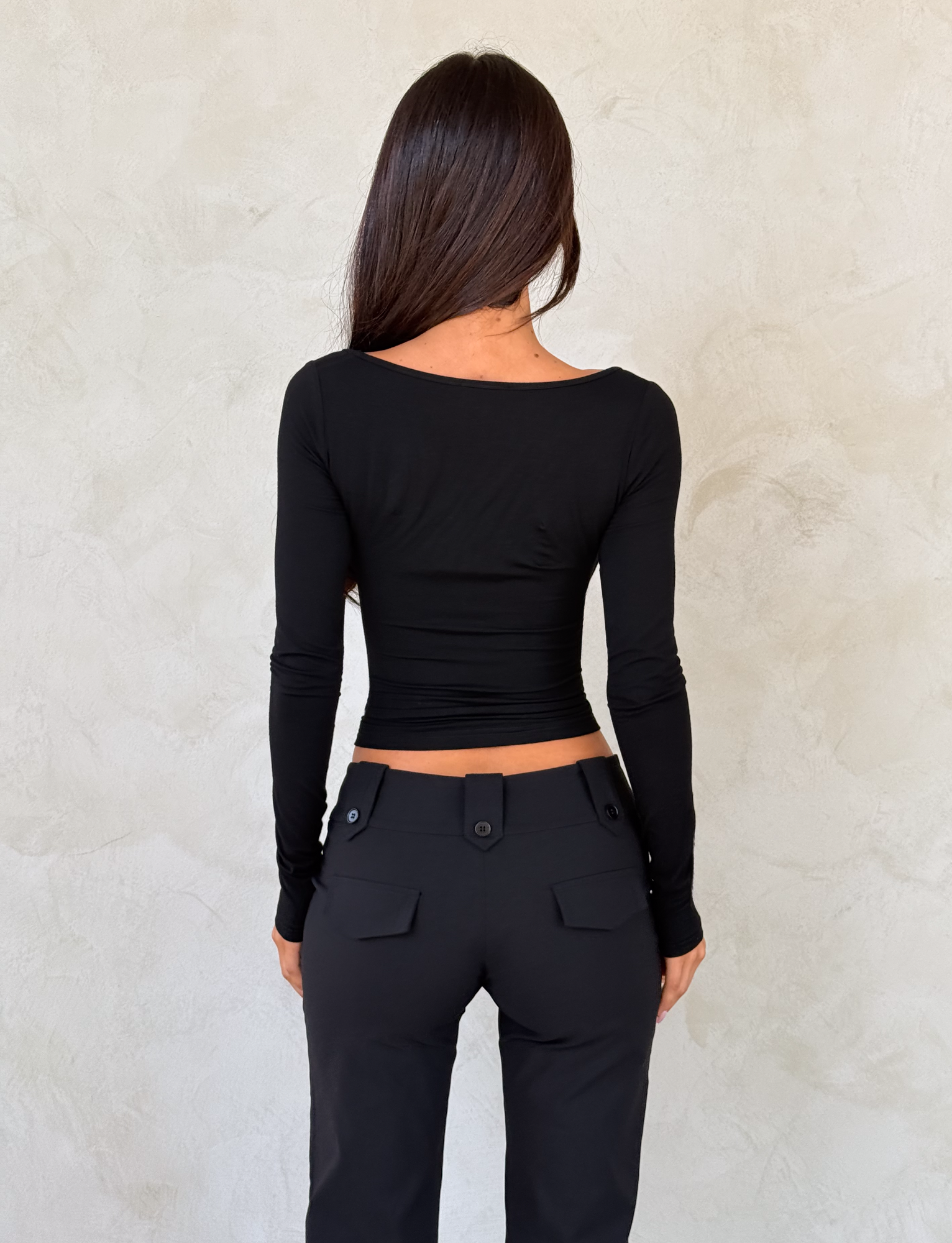 A woman with long brown hair stands facing a light wall, her back to the camera, wearing the TIGERMIST DAMELA LONG SLEEVE TOP in black and black pants with buttoned back pockets.