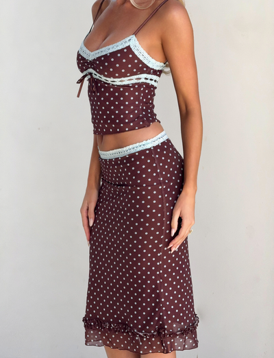 A woman wears the TIGERMIST AMARA MIDI SKIRT - POLKA DOT with a matching brown, polka-dot camisole featuring light blue lace trim, spaghetti straps, and ruffle hem for a fitted, sheer look.