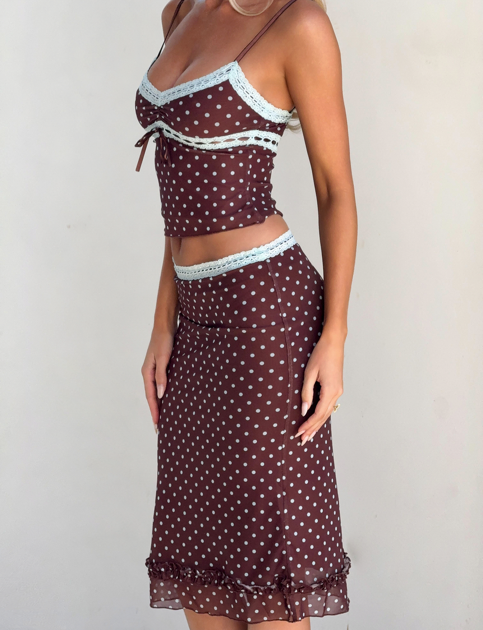 A woman wears the TIGERMIST AMARA MIDI SKIRT - POLKA DOT with a matching brown, polka-dot camisole featuring light blue lace trim, spaghetti straps, and ruffle hem for a fitted, sheer look.