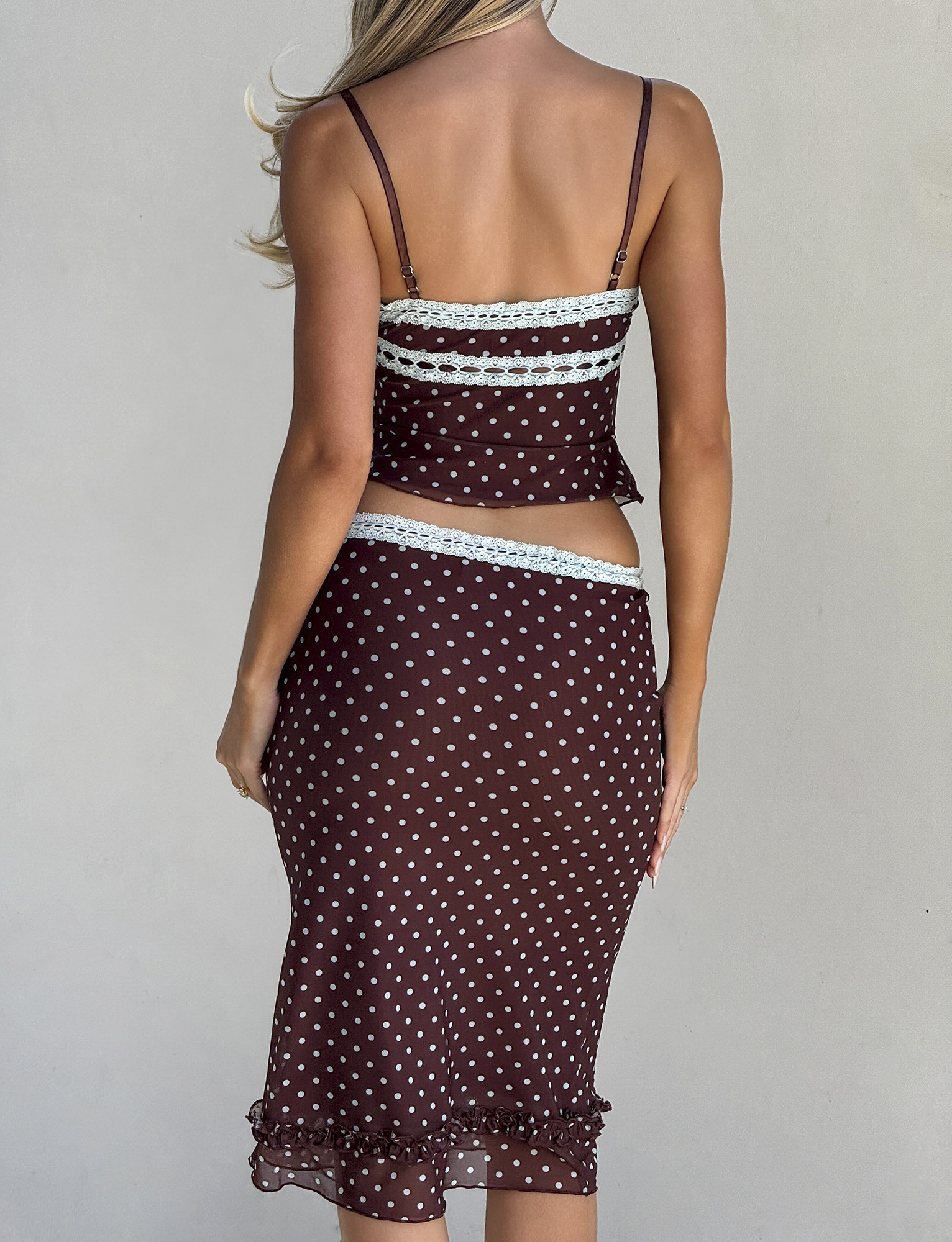 A woman with long blonde hair is seen from the back wearing the TIGERMIST AMARA TOP - POLKA DOT, a sheer brown cami with white dots, lace trim, and ruffle hem details for a feminine touch.
