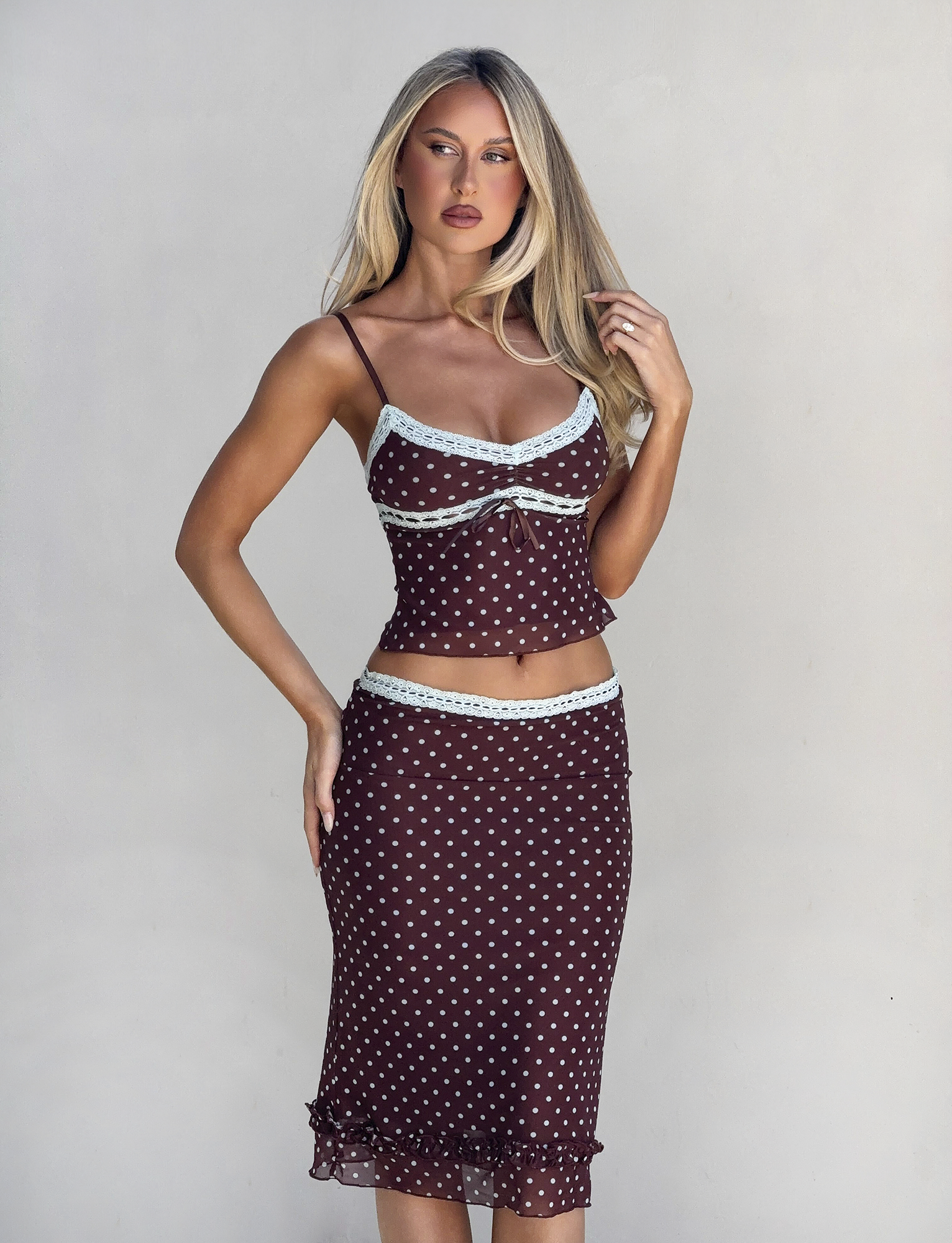 A woman with long blonde hair wears the TIGERMIST AMARA MIDI SKIRT - POLKA DOT, a brown two-piece outfit with light blue lace trim and a fitted camisole, posing against a plain light background.