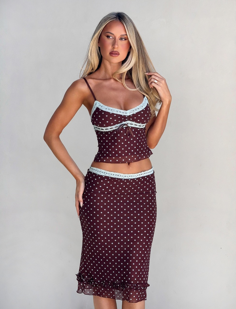 A woman with long blonde hair wears the TIGERMIST AMARA MIDI SKIRT - POLKA DOT, a brown two-piece outfit with light blue lace trim and a fitted camisole, posing against a plain light background.