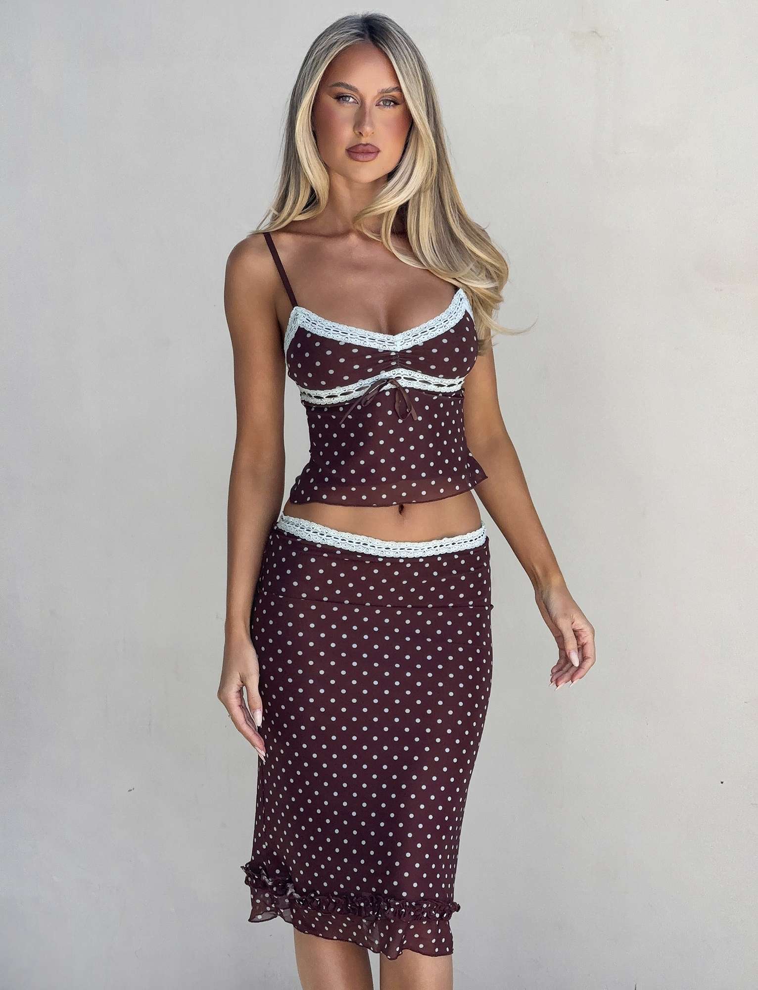 A woman with long blonde hair wears the TIGERMIST AMARA MIDI SKIRT - POLKA DOT set, featuring a fitted cami top and a ruffled-hem midi skirt with light blue lace trim, standing against a plain light background.