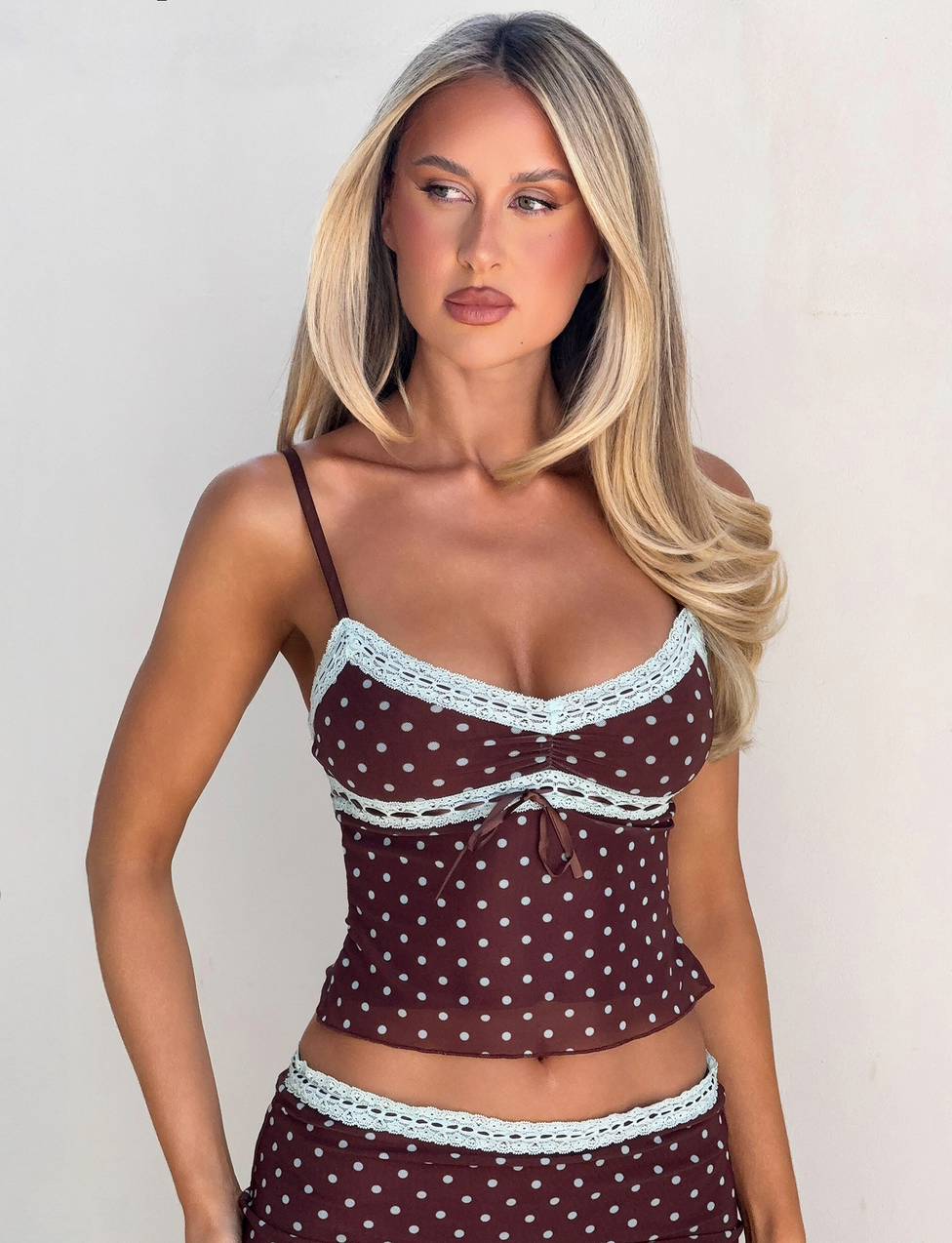 A woman with long blonde hair models the TIGERMIST AMARA TOP - POLKA DOT, a brown cami with light blue lace trim, against a plain light background, gazing to the side with a neutral expression.