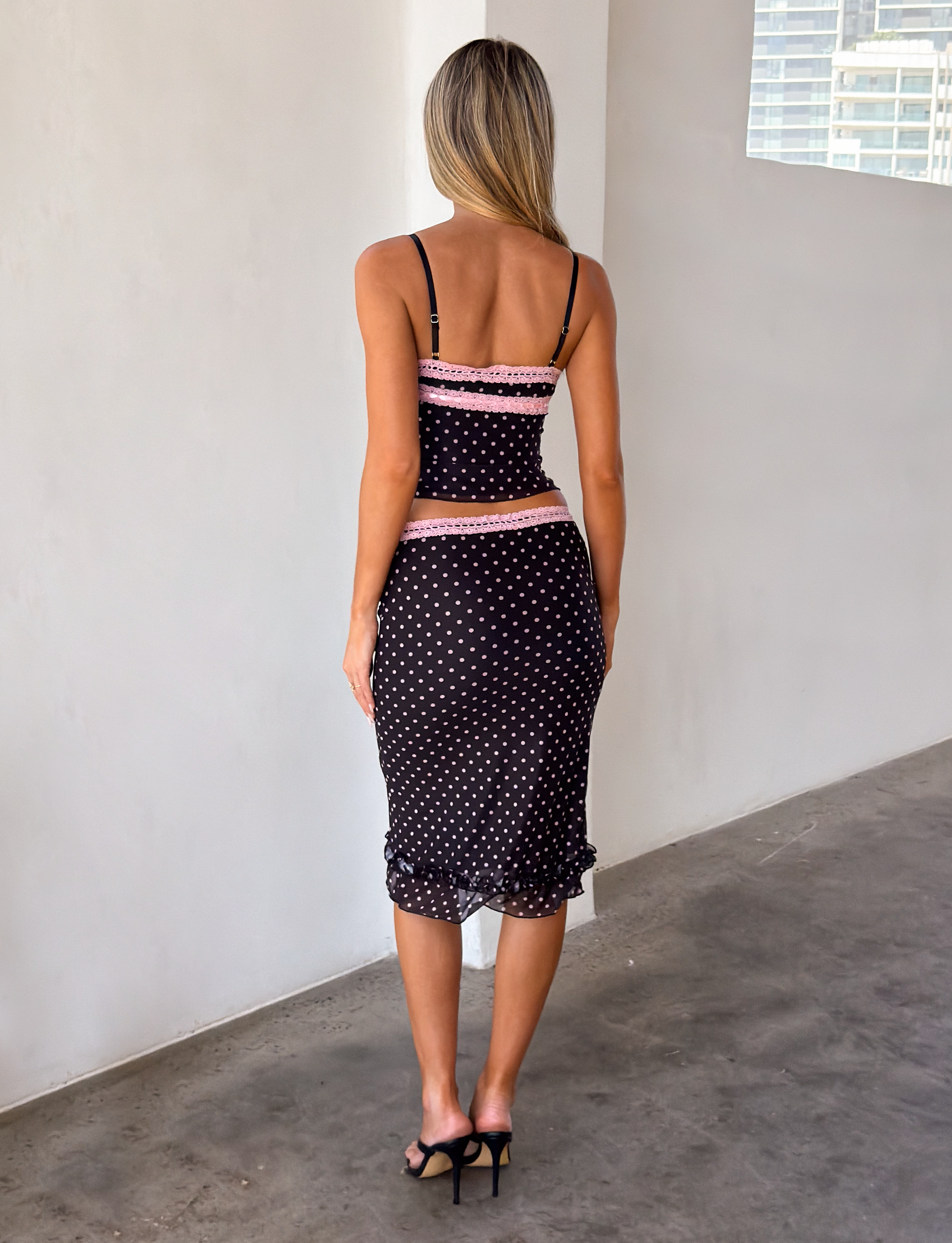 A woman with long blonde hair stands indoors facing away, wearing the TIGERMIST AMARA TOP - BLACK, paired with a polka dot cami dress and black high-heeled sandals. The space has white walls and a concrete floor.