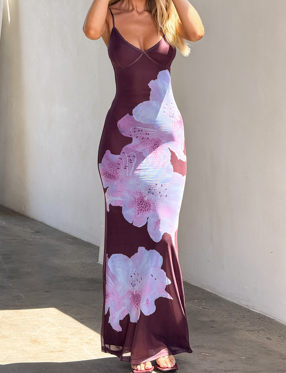 A woman in the TIGERMIST ORIELLE MAXI DRESS - PLUM ORCHID, a fitted, sleeveless plum maxi with thin straps and a bold floral print, poses against a white wall, hands gently touching her hair.