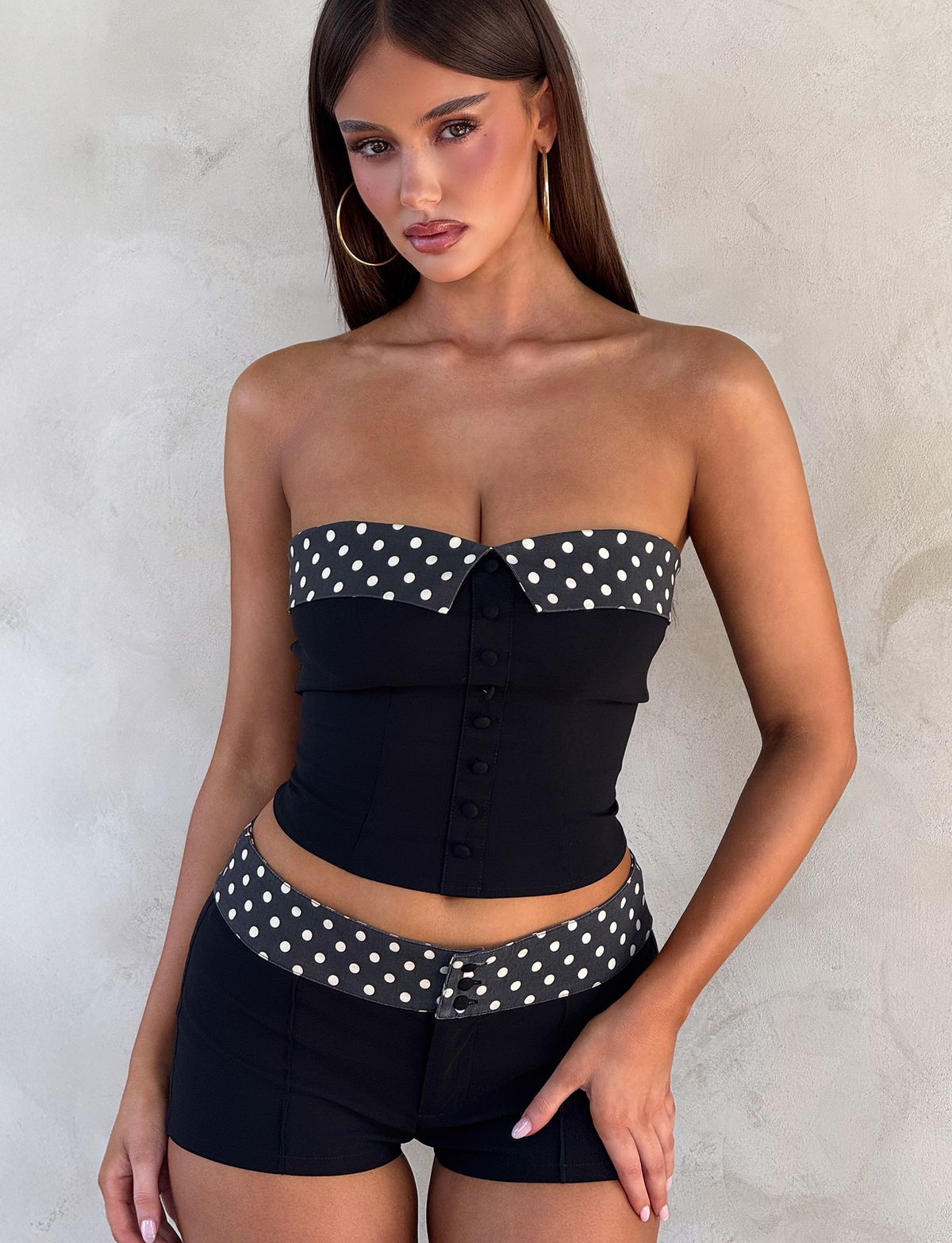 A woman with long straight hair and large hoop earrings stands against a light textured wall, wearing the TIGERMIST KITTIE 2.0 BUSTIER in black, paired with matching shorts featuring black and white polka-dot trim.