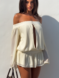 A woman poses against a white background wearing the TIGERMIST TESSIE PLAYSUIT in ivory—an off-shoulder design featuring long sleeves, a front slit, and a ruffled skirt—while holding a handbag.