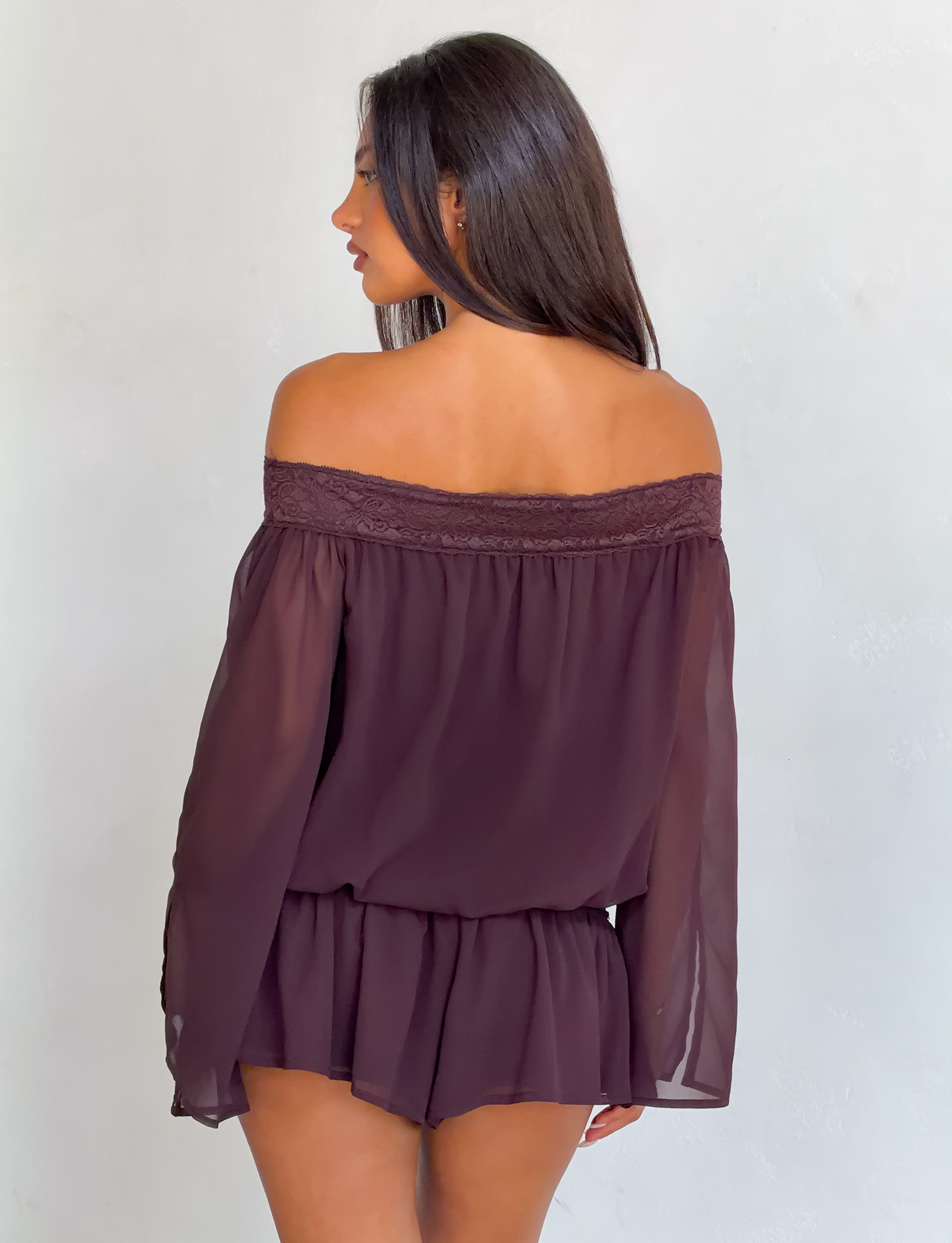 A woman with long dark hair faces away, wearing the TIGERMIST TESSIE PLAYSUIT in brown. The off-shoulder design features sheer fabric, lace trim, and ruffled mini shorts for a loose, flowy fit. The background is plain and light.