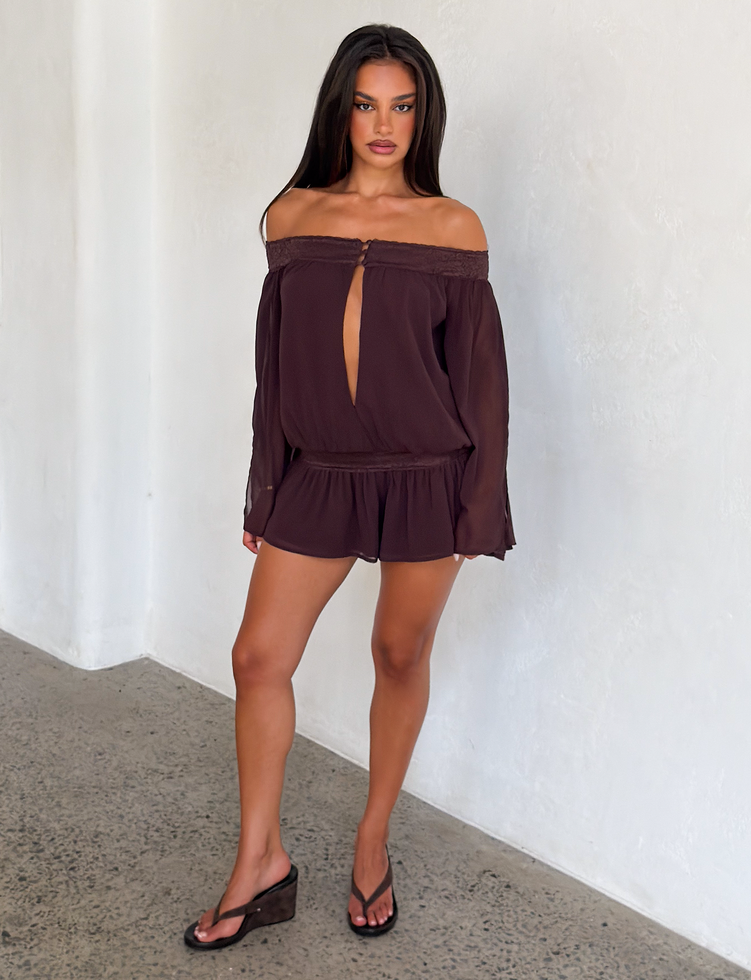 A woman with long dark hair and a neutral expression stands indoors against a light textured wall, wearing the TIGERMIST TESSIE PLAYSUIT in brown—an off-shoulder style featuring ruffled mini shorts, long sleeves, and a front keyhole cutout—paired with black sandals.