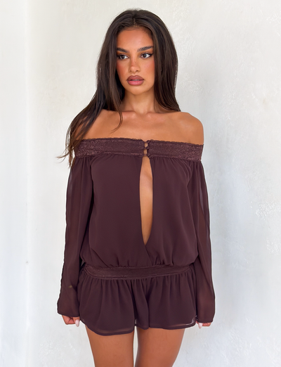 A woman models the TIGERMIST TESSIE PLAYSUIT in brown, featuring an off-shoulder design with long sleeves and ruffled mini shorts, standing against a plain white textured wall and looking slightly away from the camera.