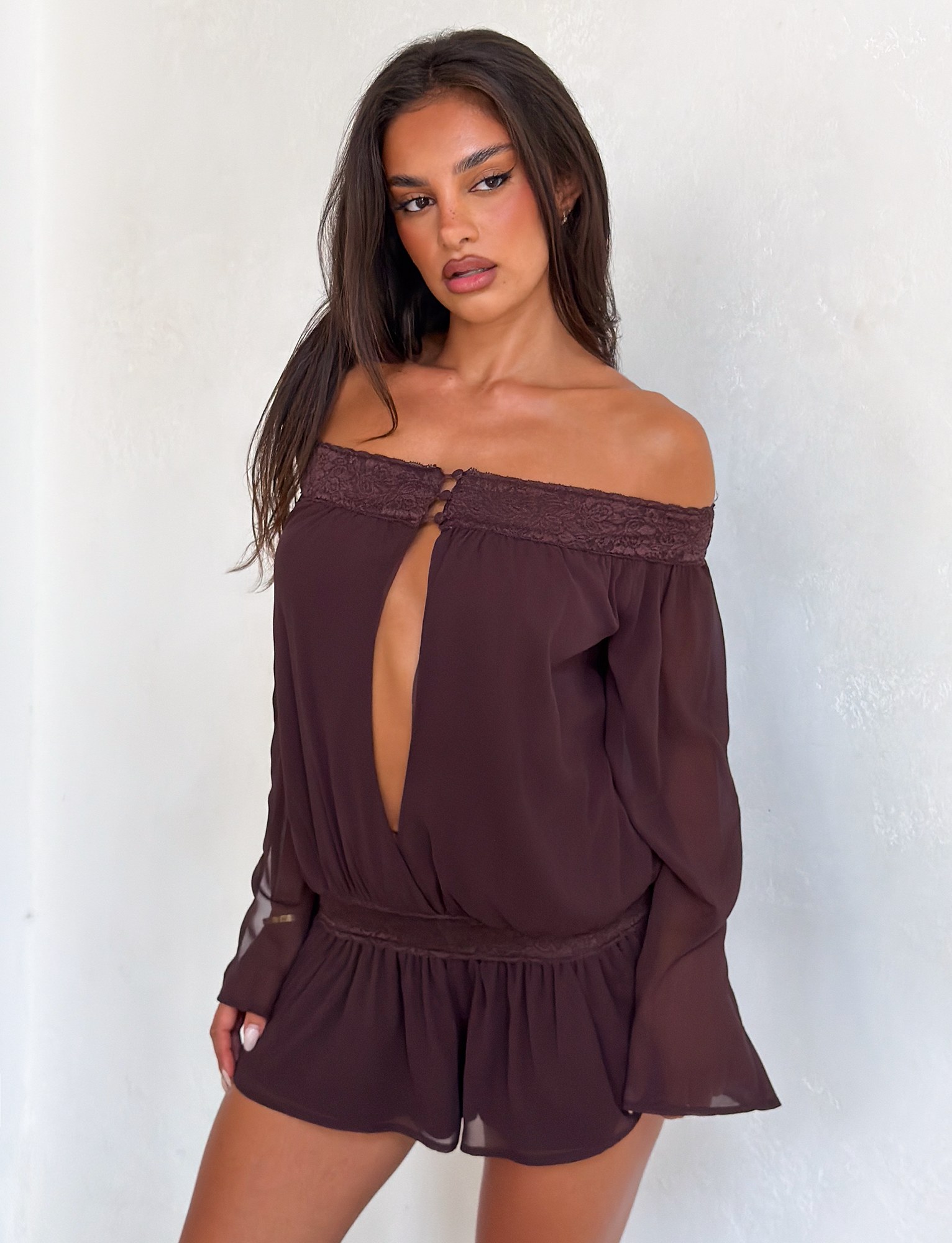 A woman with long dark hair wears the TESSIE PLAYSUIT in brown by TIGERMIST, featuring an off-the-shoulder design, front cutout, and ruffled mini shorts, standing against a plain white wall.