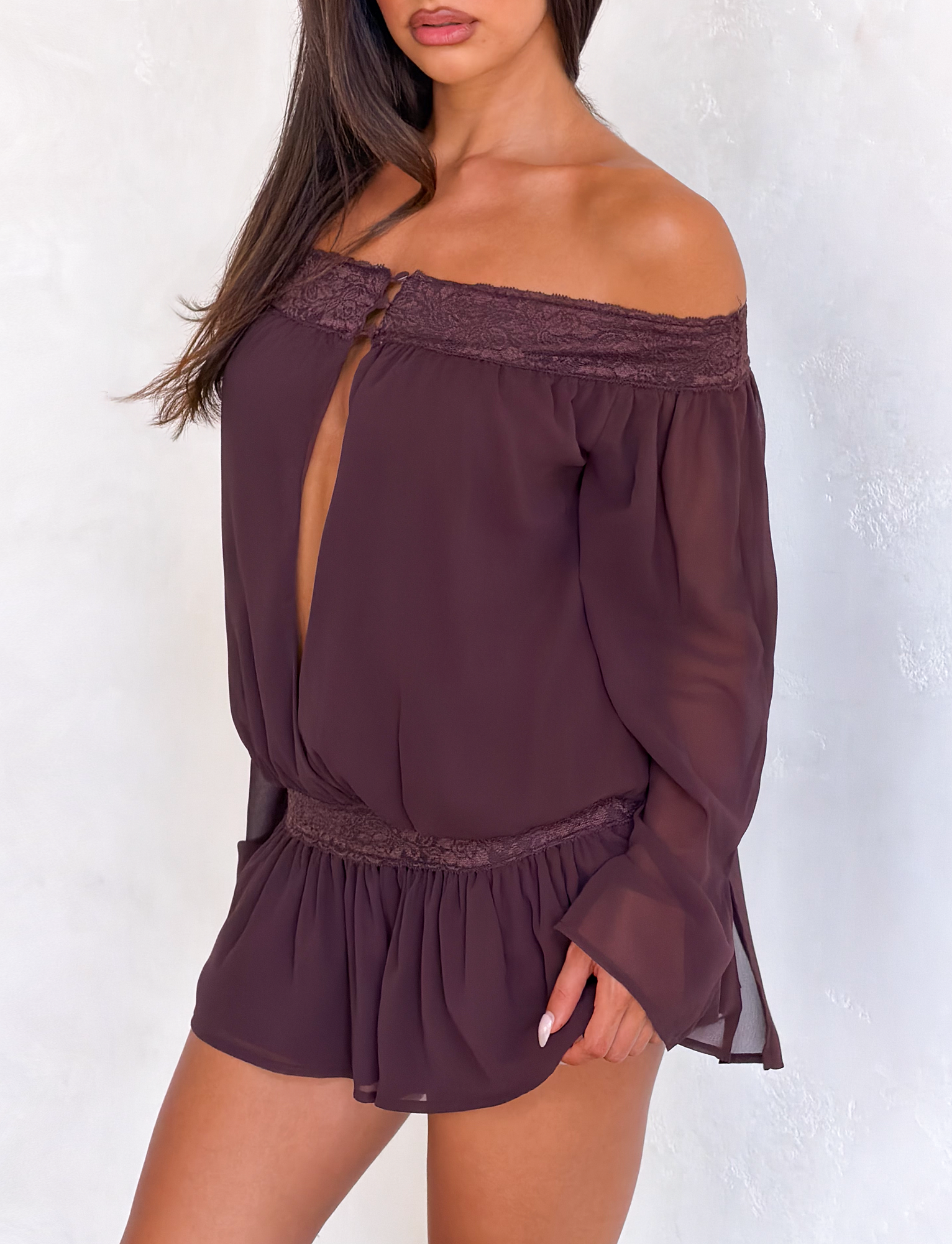 A woman models the TIGERMIST TESSIE PLAYSUIT in brown, a sheer, off-shoulder style with lace trim at the neckline and waist, and ruffled mini shorts. She stands against a light background, her face partially visible.