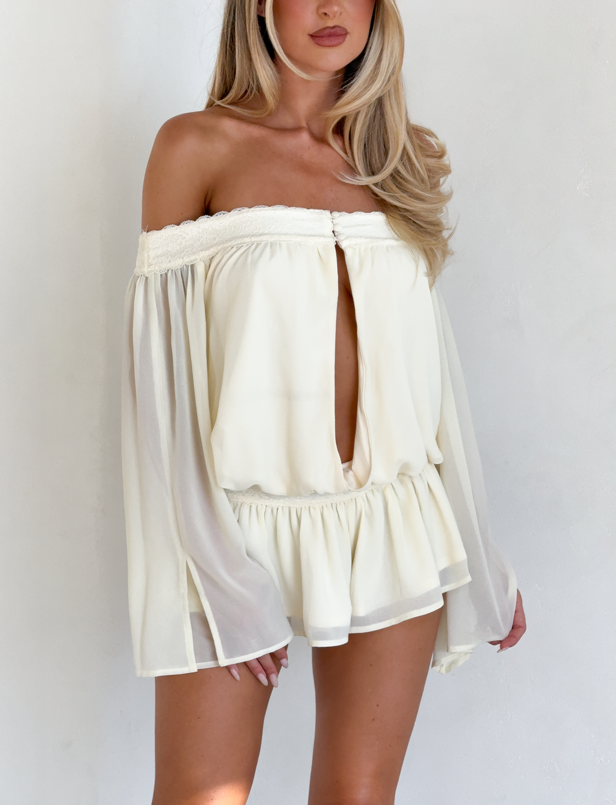 A woman stands against a plain white background wearing the TIGERMIST TESSIE PLAYSUIT in ivory, featuring an off-shoulder design, ruffled mini shorts, and a front keyhole cutout. Her blonde hair falls over her shoulders.