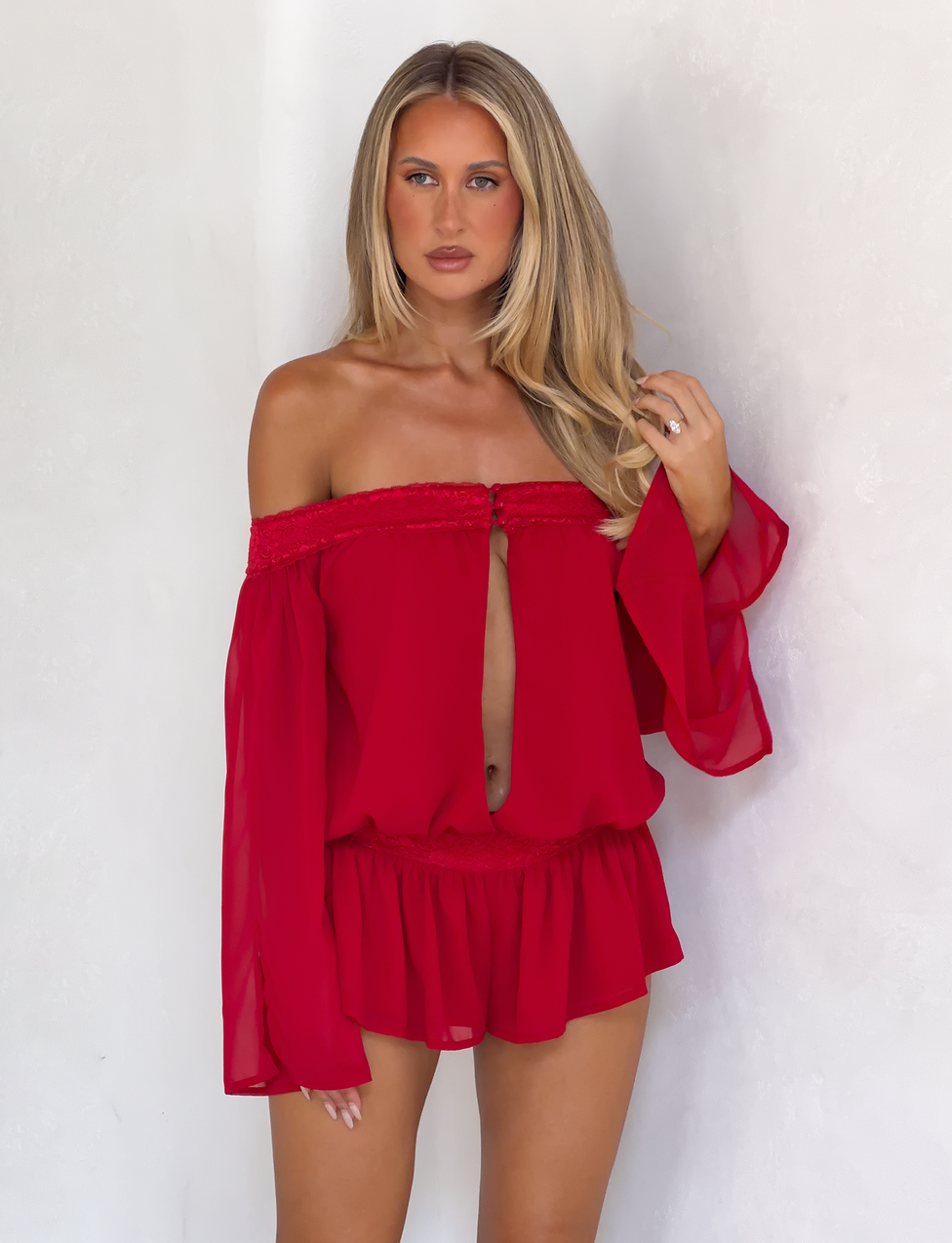 A woman poses against a white background in the Tiger Mist USA CL - TESSIE PLAYSUITS, a bright red, off-the-shoulder romper featuring long flowy sleeves and a front cutout. Her light hair is styled down as she looks at the camera.
