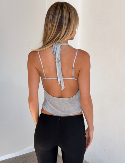 A woman with long blonde hair faces away, wearing the TIGERMIST MAKENA TWO-PIECE TOP 2.0 in silver and black pants, standing in a minimalist, light-colored room.