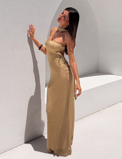A woman models the TIGERMIST DANIELA SCARF DRESS 2.0 - GOLD, posing against a white, curved backdrop with one hand on the wall, head tilted back and eyes closed, bathed in bright sunlight.
