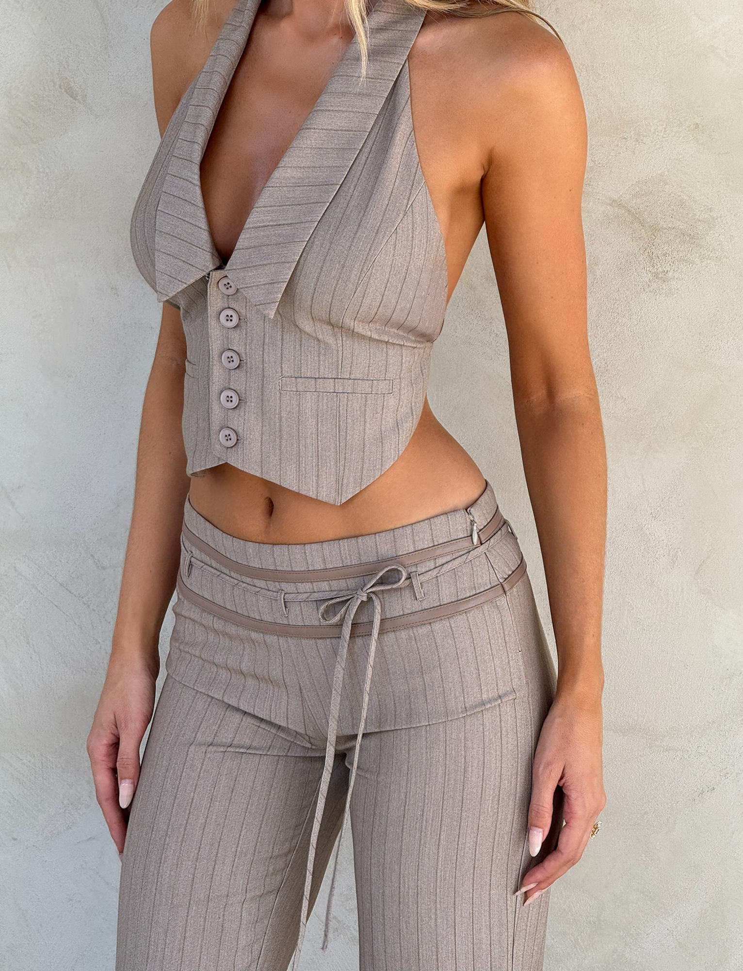 A woman models the TIGERMIST FEDRA VEST - TAN PINSTRIPE with matching high-waisted trousers and tie belt, standing against a light textured wall. Her face is turned away from the camera.