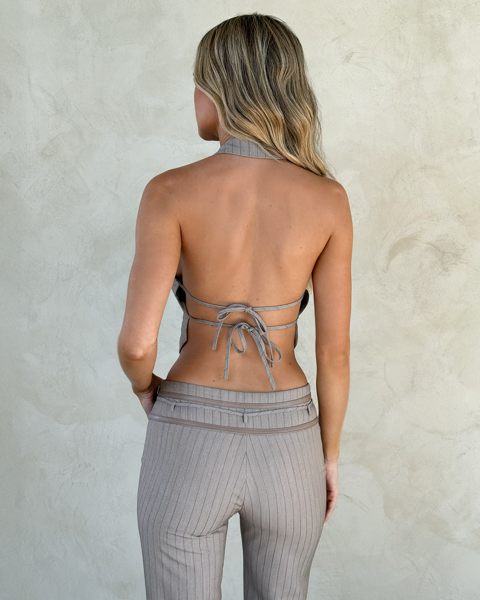 A woman with long, wavy blonde hair wearing TIGERMIST’s FEDRA PANT in tan pinstripe stands facing a light textured wall. She pairs the pants with a backless halter top, her back visible and arms relaxed at her sides.